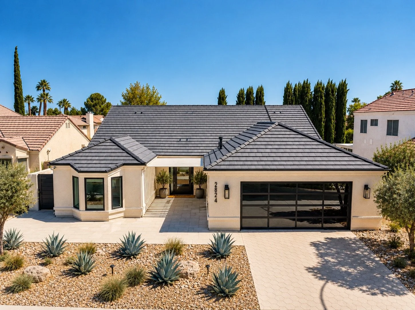 Front view of a modern single-story house with a dark gray tile roof, beige exterior walls, large windows, and a two-car garage, with desert landscaping including agave plants and small shrubs, under a clear blue sky.
