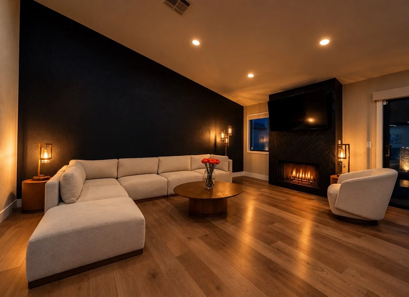 Cozy living room with white sectional sofa, armchair, round wooden coffee table with flowers, fireplace, black accent wall, ceiling lights, side lamps, and a window with a view of the night outside.