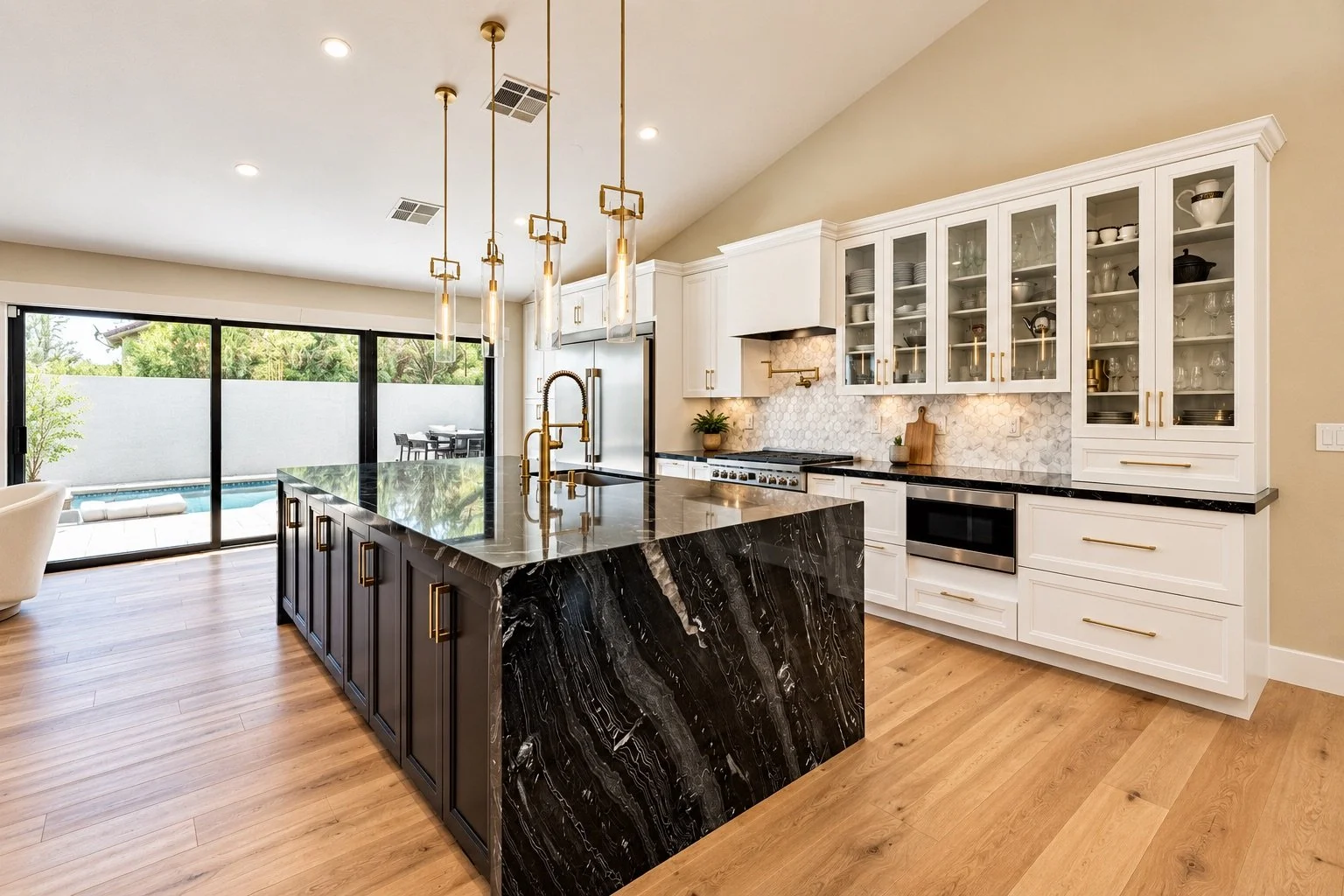 Modern kitchen with white cabinets, a black marble island with gold hardware, glass-front cabinets, and wood flooring, overlooking a backyard with a pool.