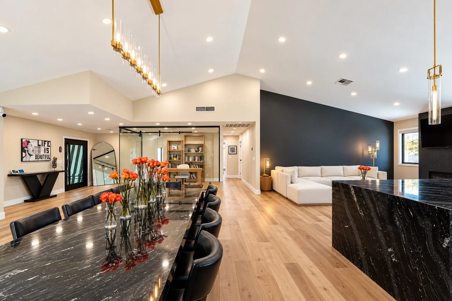 Modern open-concept living room and dining area with black marble table, white sectional sofa, and black accent wall, decorated with orange flowers and gold lighting fixtures.