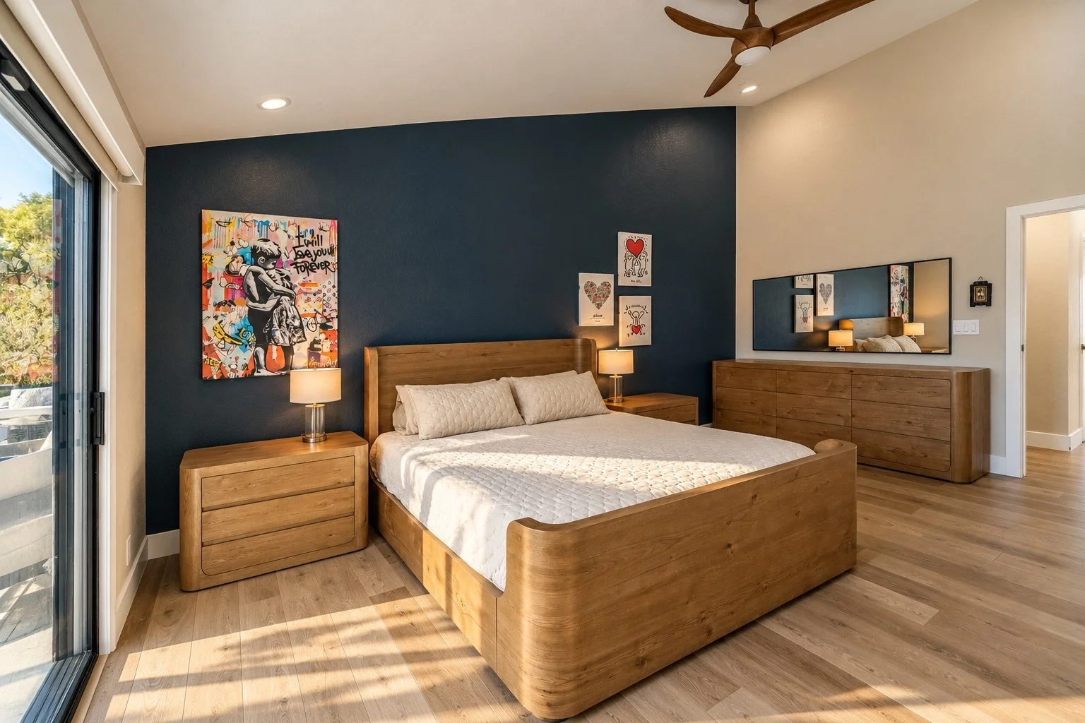 Modern bedroom with a wooden bed frame, white bedding, and matching side tables. Contains colorful artwork and a large mirror on the wall. The room has hardwood flooring and a ceiling fan. A sliding glass door opens to an outdoor balcony with scenic views.
