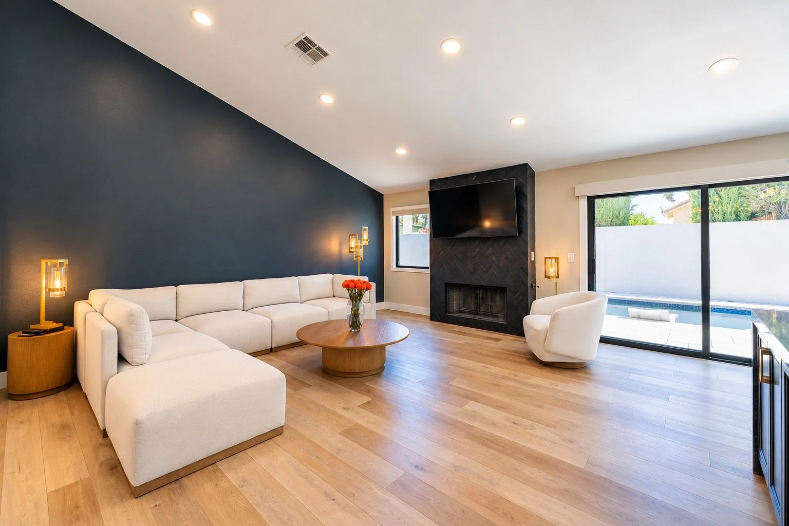 Modern living room with a white sectional sofa, a round wooden coffee table with a vase of flowers, a white armchair, a black accent wall with a wall-mounted TV above a fireplace, and sliding glass doors leading to a backyard pool area.