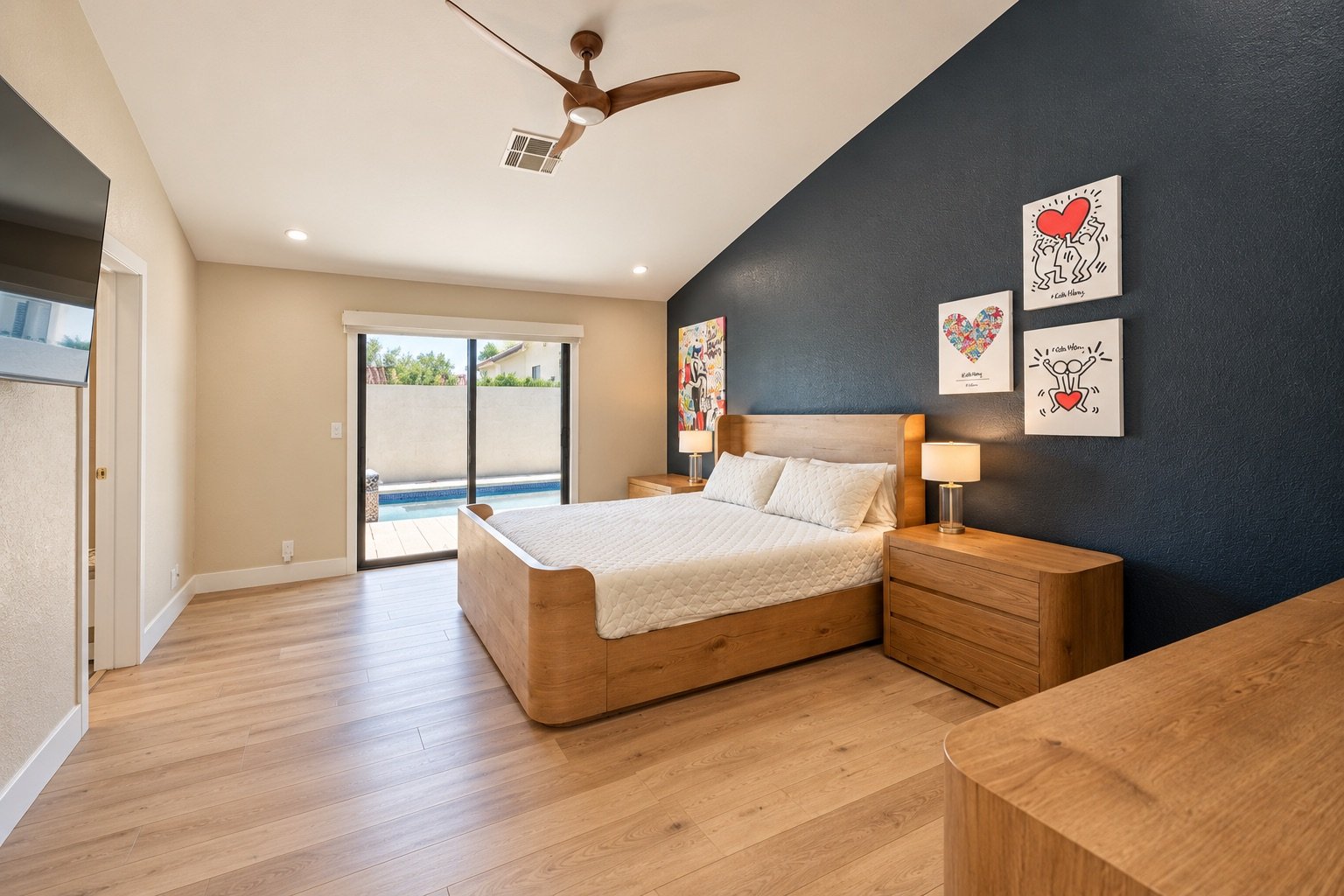 Bedroom with wooden bed frame, white quilt, matching pillows, two wooden nightstands with lamps, artwork on dark blue wall, and sliding glass door leading to backyard pool area.