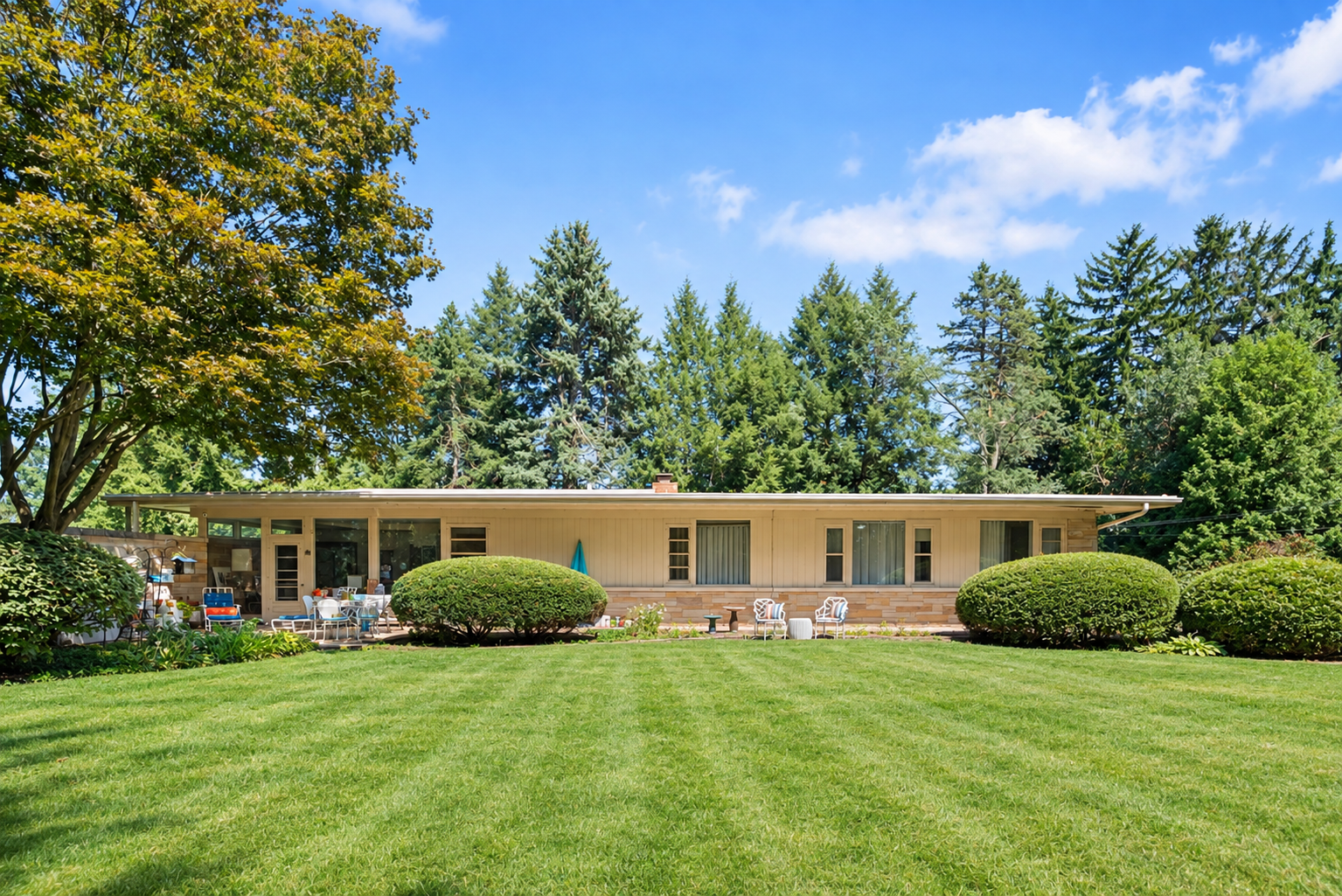 19 Greenacres backyard view of a single-story house with a large patio, surrounded by one acre of lawn, shrubs, and mature trees.