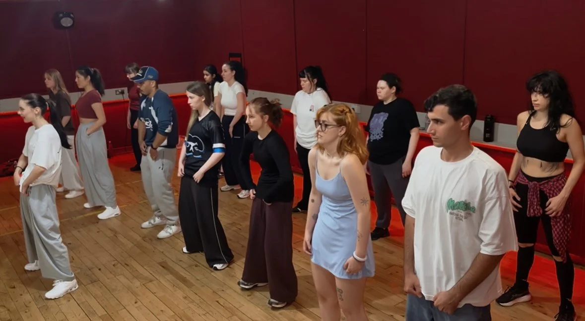 Group of people in a dance studio practicing dance moves, standing in rows on a wooden floor against a red wall. Dublin dance basics class, dublin dance fundamental class