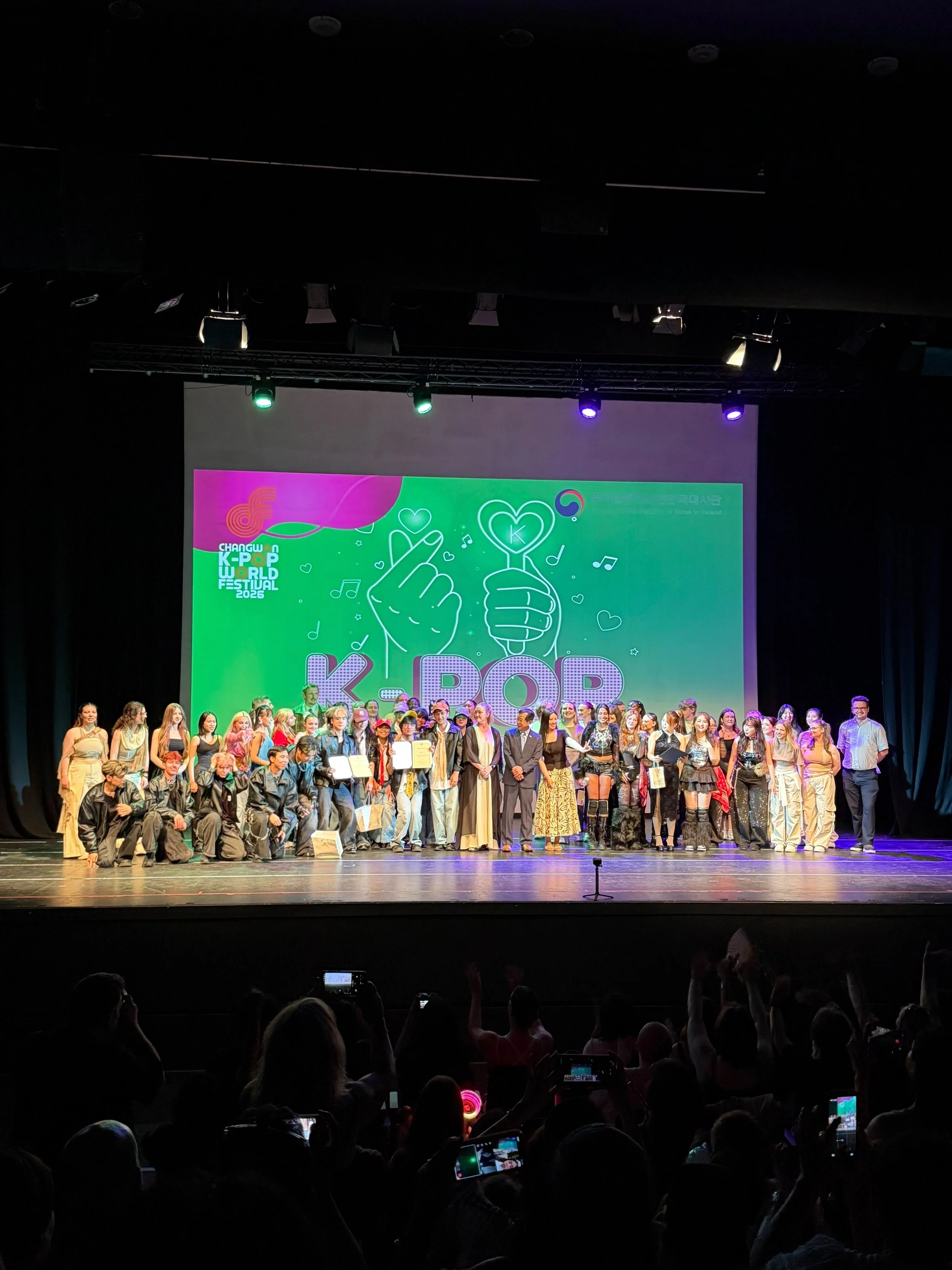 Ireland Dublin K-pop World Festival, with many holding certificates or awards, and audience members taking photos. Judge Korean Rosaile Han
