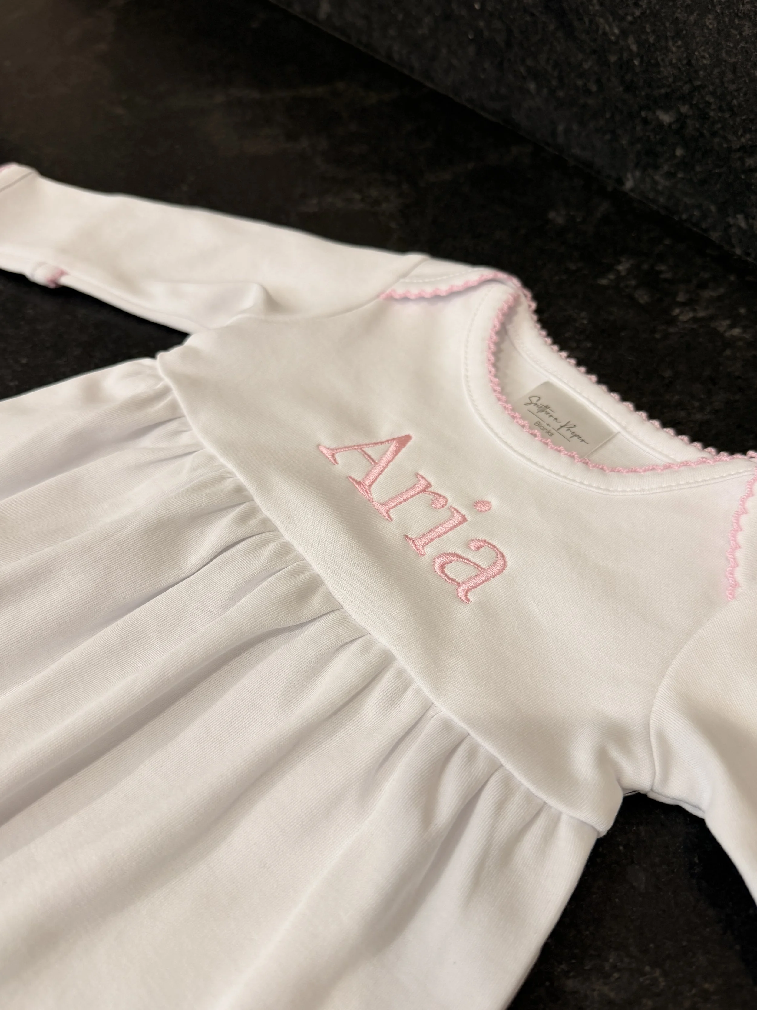 A white baby dress with pink embroidery and ruffled skirt laid on a black surface.
