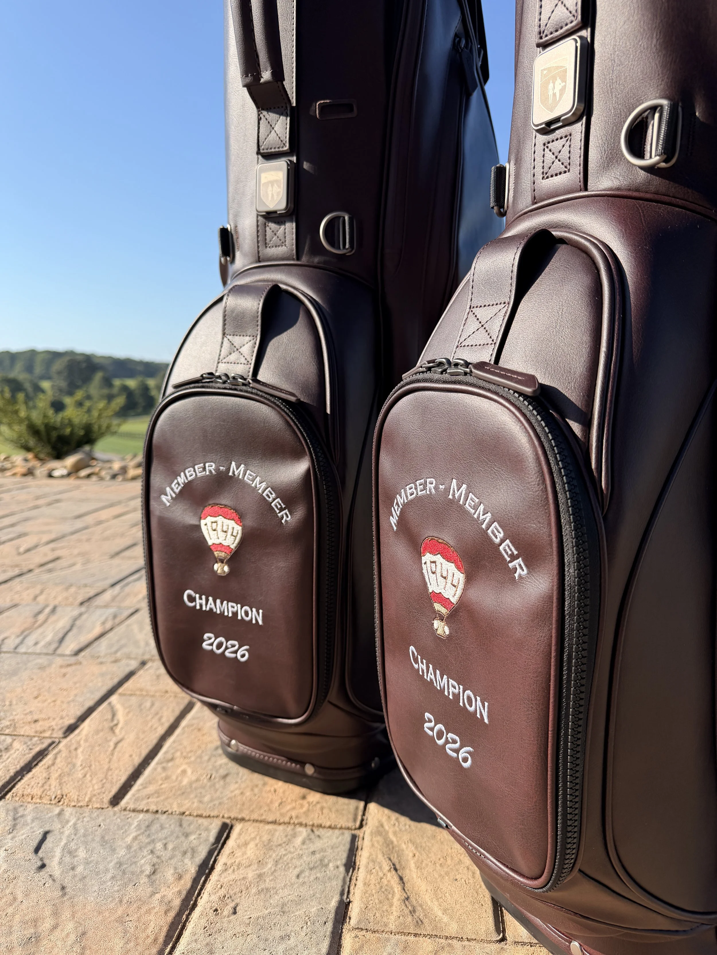 Two brown golf bags with zippers and a logo that says 'Member-Member Champion 2026' are standing on a stone pathway outdoors with a blurred background of greenery and a clear blue sky.