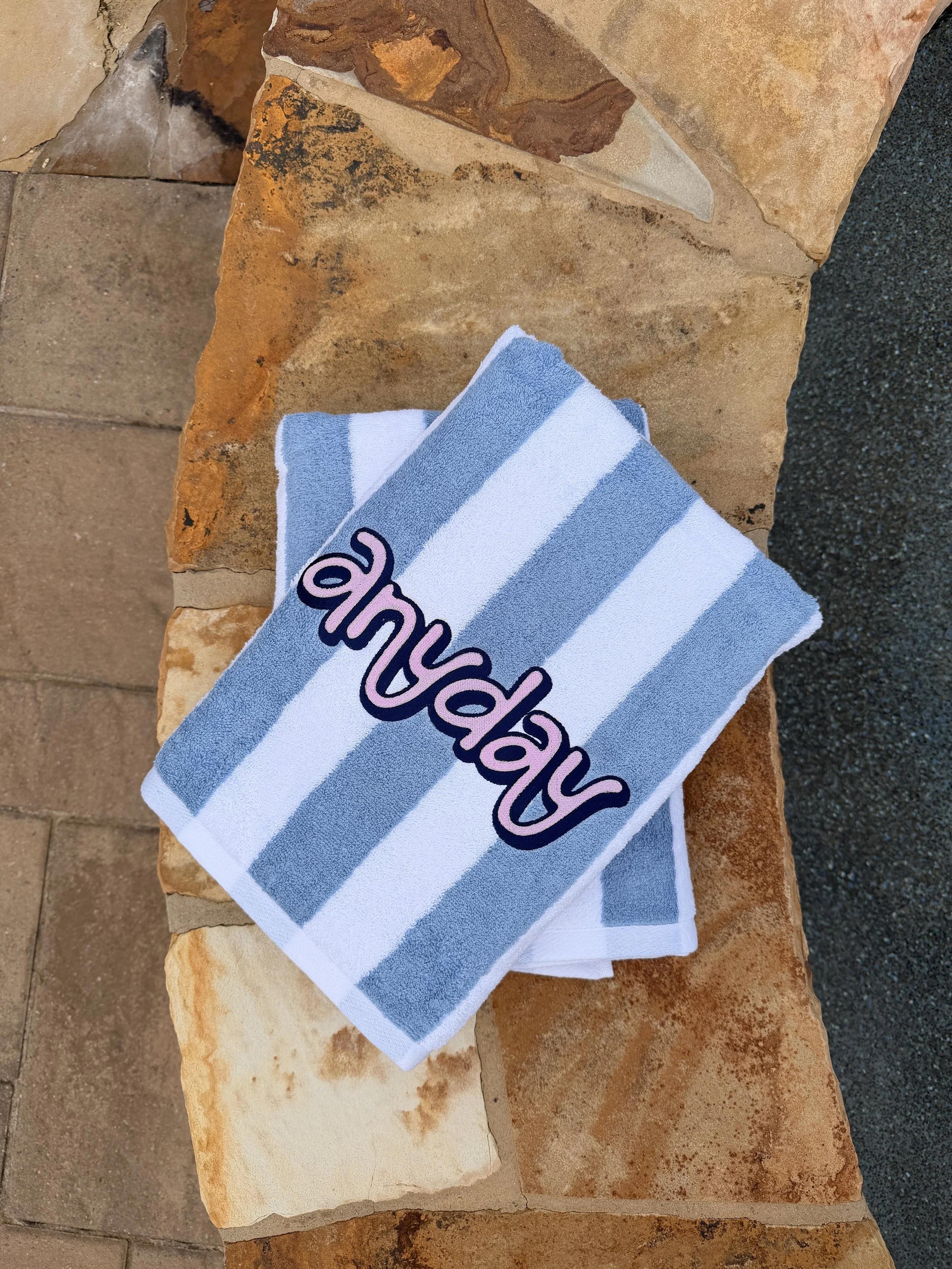 A towel with blue and white horizontal stripes and purple text, placed on a stone surface with a brick pavement in the background.