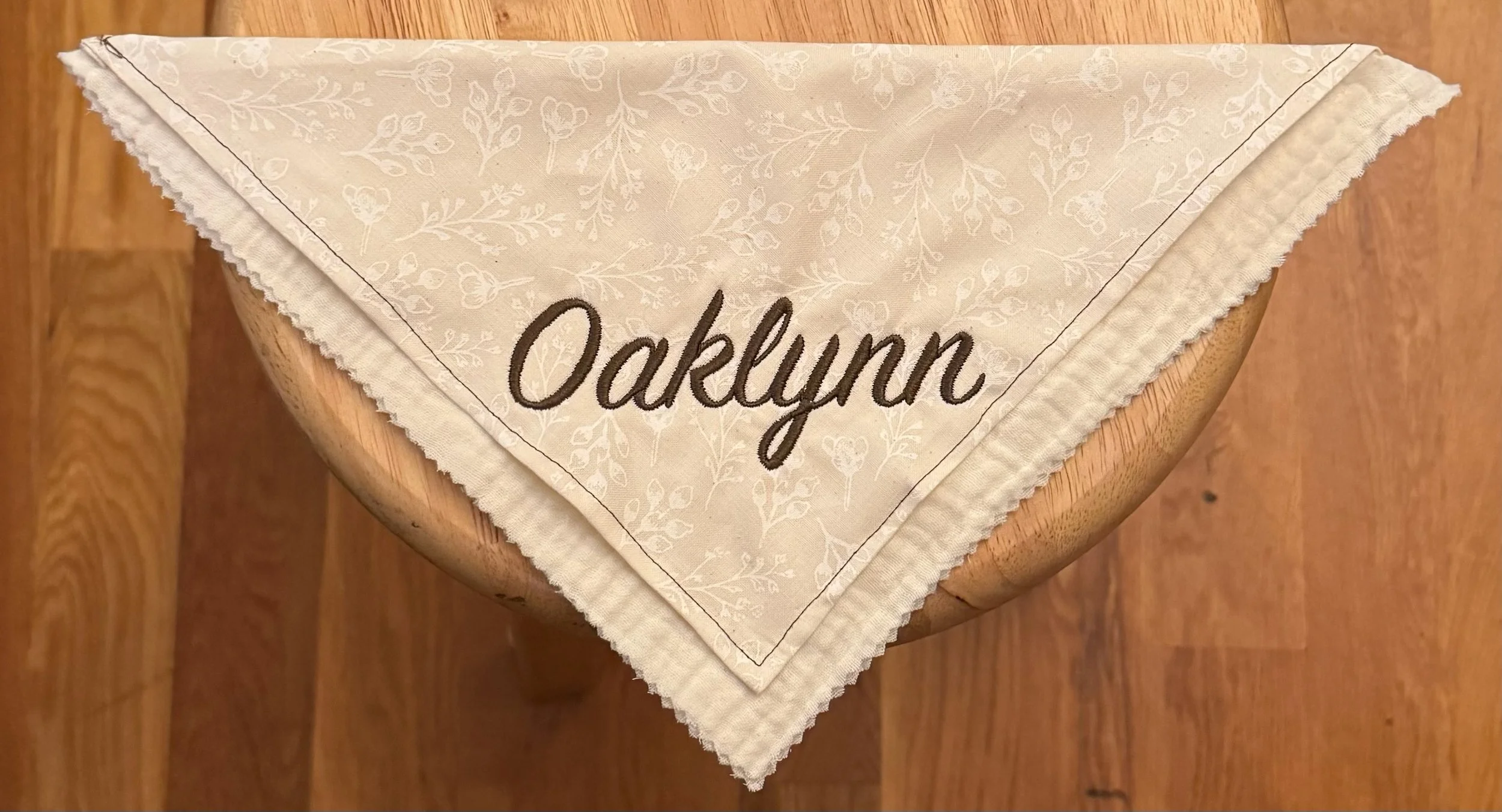 A folded white cloth napkin with a floral pattern and the name 'Okayun' embroidered in dark brown, placed on a wooden chair.