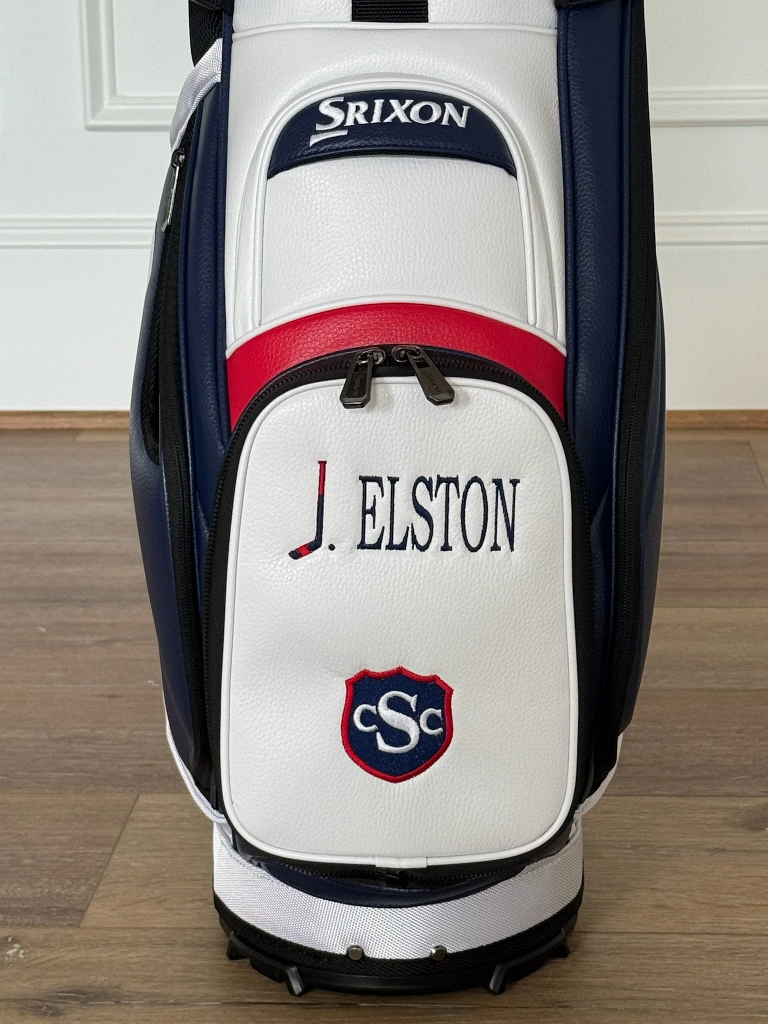 A white golf bag with red and navy accents, featuring the name 'J. Ellston' and an SC logo patch, standing on a wooden floor against a white paneled wall.