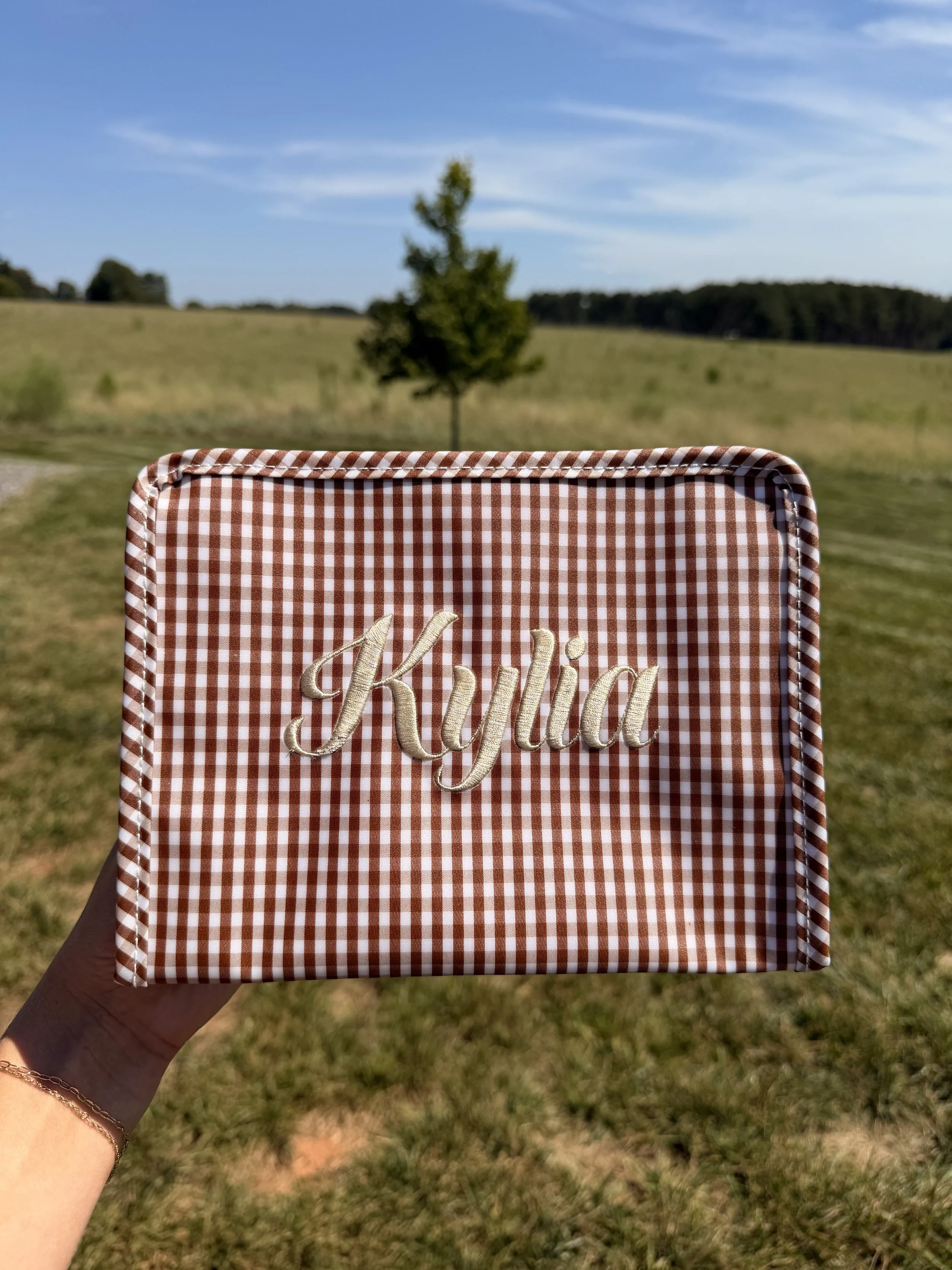Close-up of a hand holding a checkered cloth with the word "Honey" embroidered on it, outdoors on a sunny day with a grassy field and a small tree in the background.