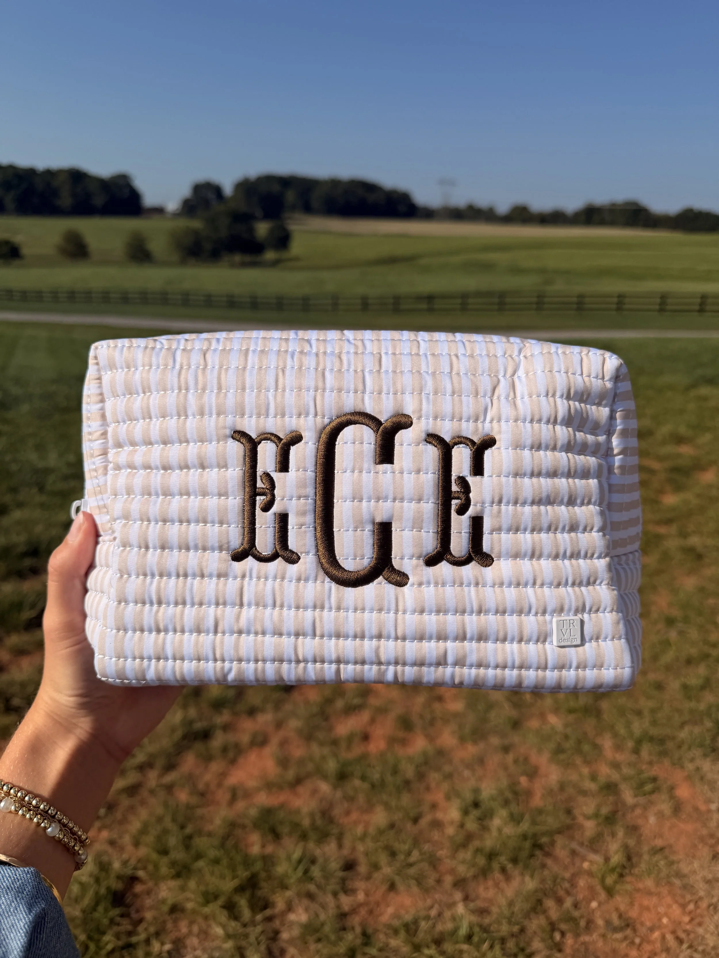 A person holding a quilted fabric item with the name 'Harlow' embroidered on it, outdoors in a grassy field with trees and a fence in the background under a clear blue sky.