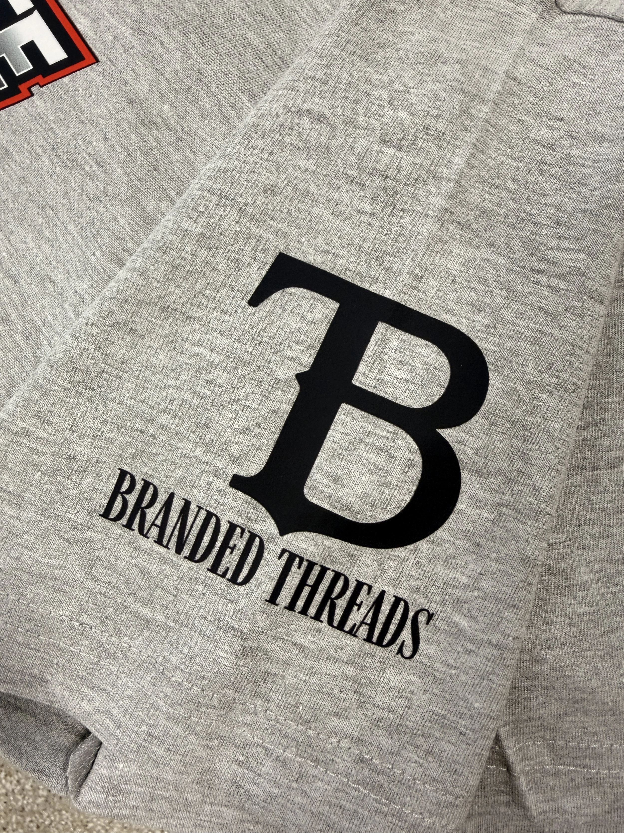 Close-up of a gray t-shirt with black text that says 'BRANDED THREADS' and a large letter 'B' in black in the center, with part of a red and white logo visible in the top left corner.