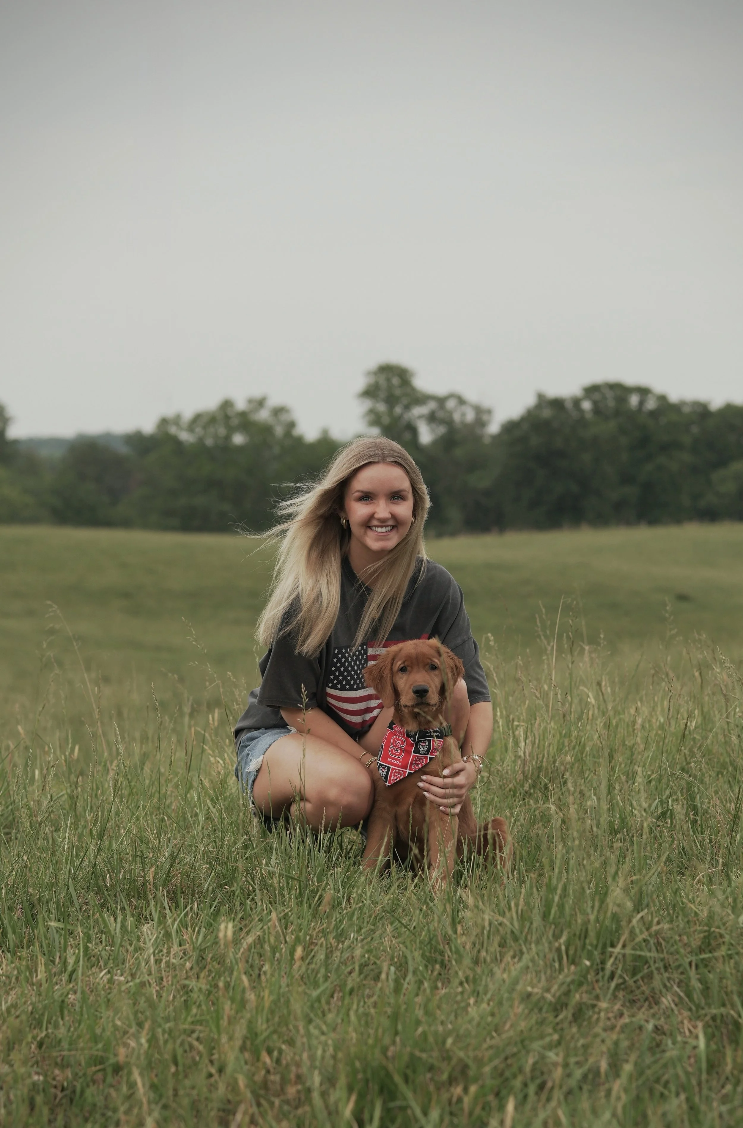 A young woman with long blonde hair smiling while crouching in a grassy field, holding a brown retriever puppy wearing a red bandana, with trees and a cloudy sky in the background.
