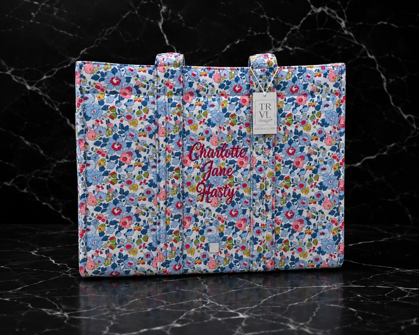 A fabric book cover with colorful floral print, embroidered with the names Charlotte, Jane, and Hasty in red, placed on a black marble surface with a black marble background.