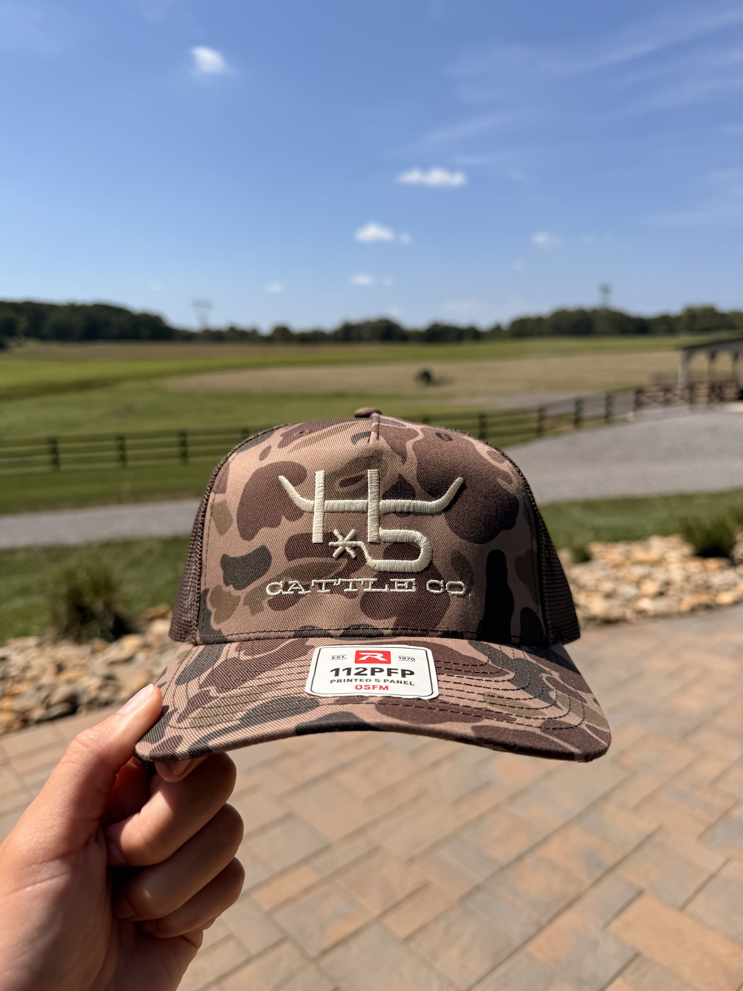 A hand holding a camo baseball cap with what appears to be a golf course and blue sky in the background.