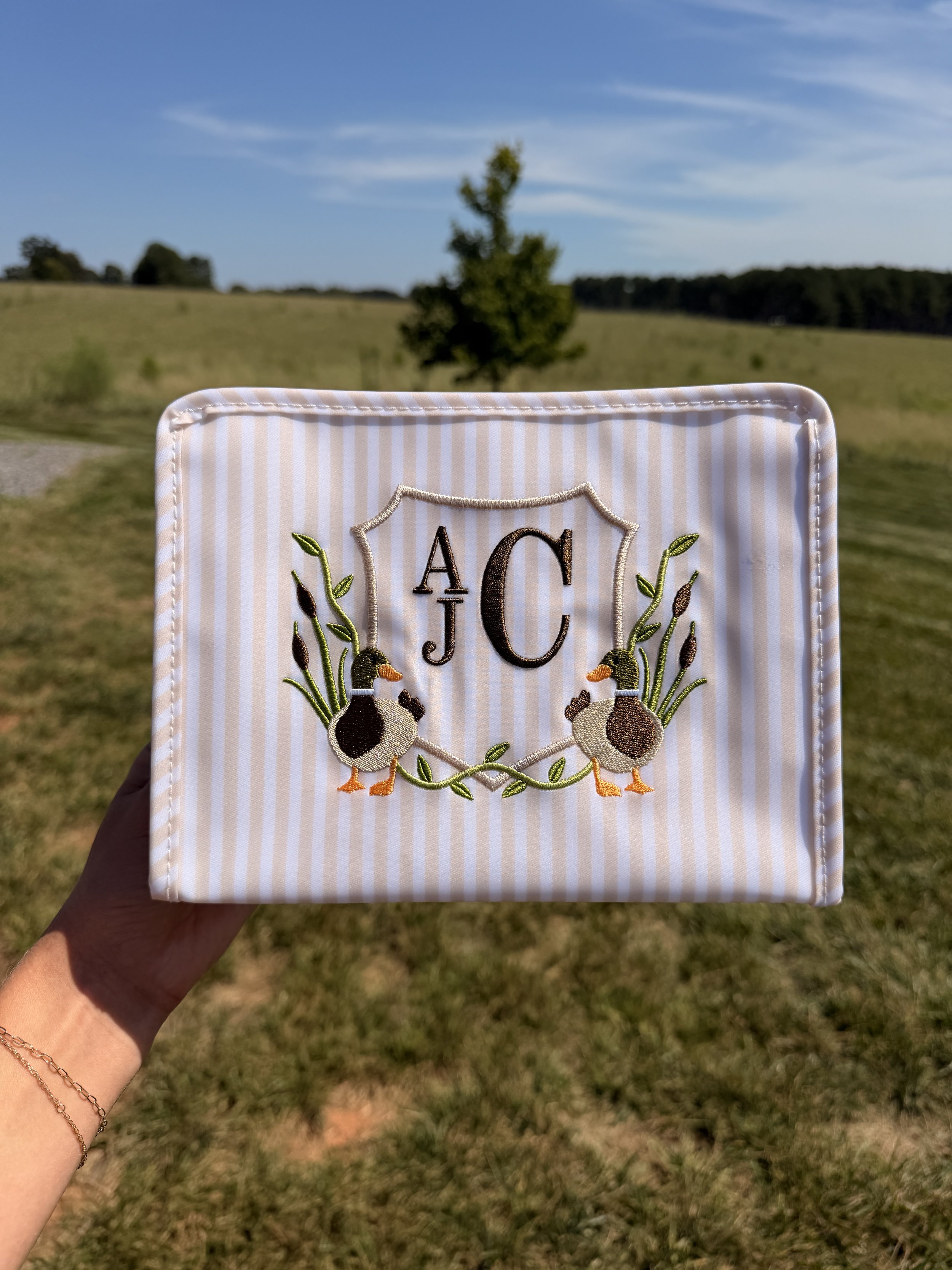 Close-up of embroidered cloth with crest featuring two ducks, foliage, and the letters 'A J C', held by a person in an outdoor field with grass, trees, and a blue sky in the background.