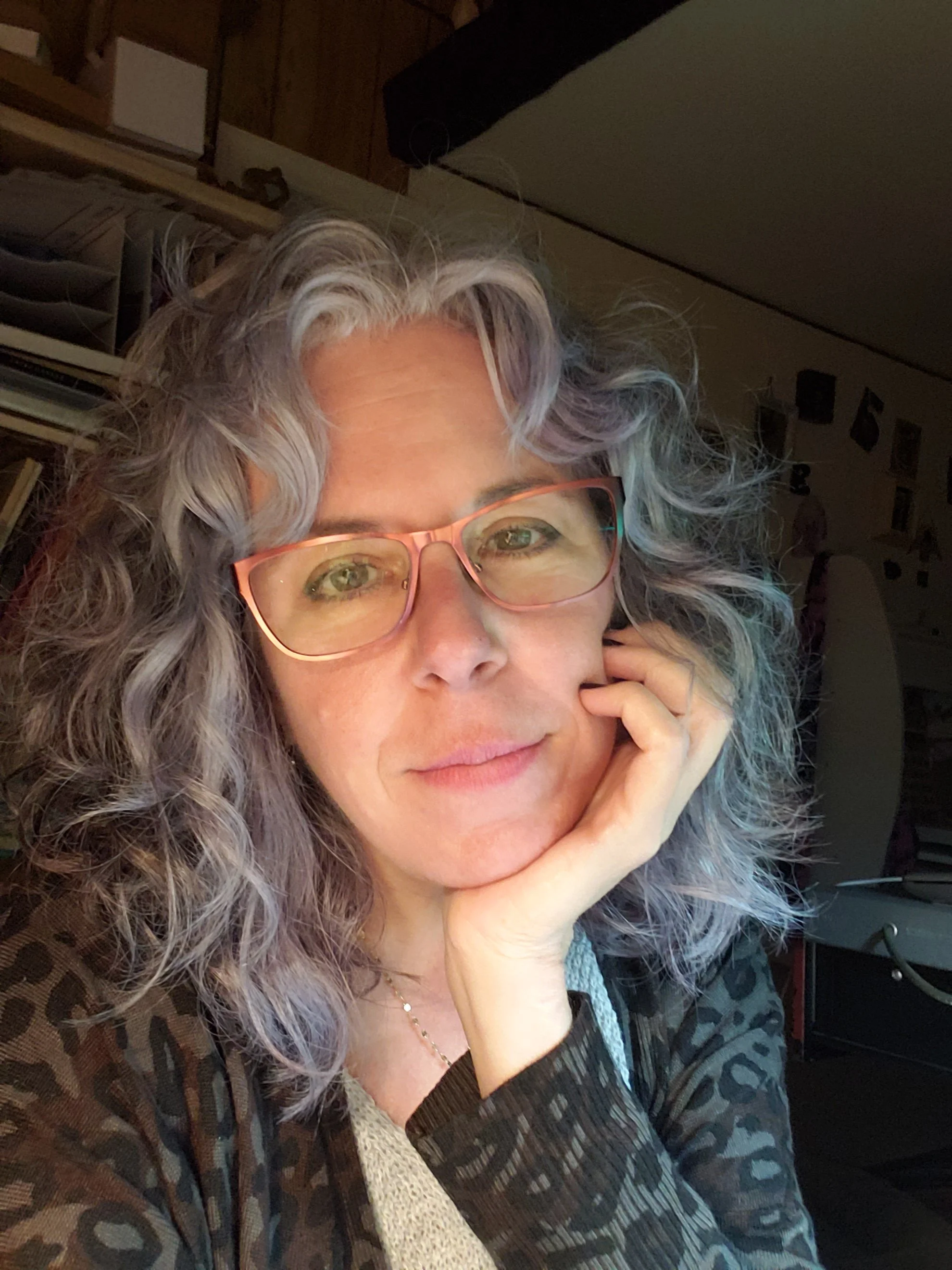 Close-up of a woman with curly gray hair, wearing glasses, resting her chin on her hand in a cozy indoor setting.