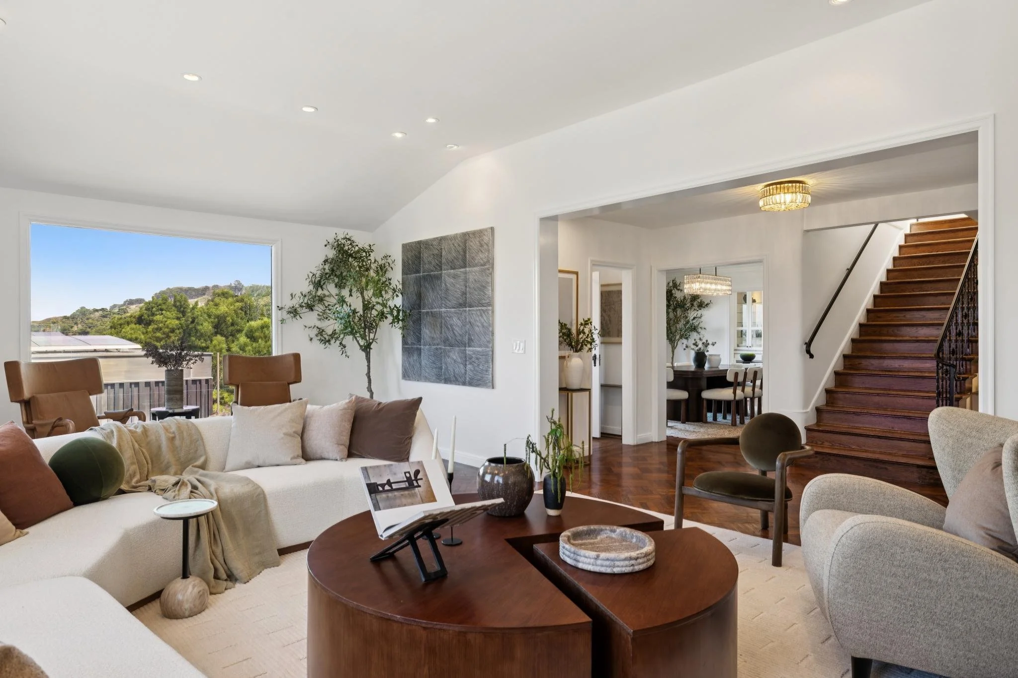 Bright, spacious living room with white walls and ceiling, hardwood floors, a large window showing green trees, surrounded by modern furniture including sofas, chairs, and decorative plants.