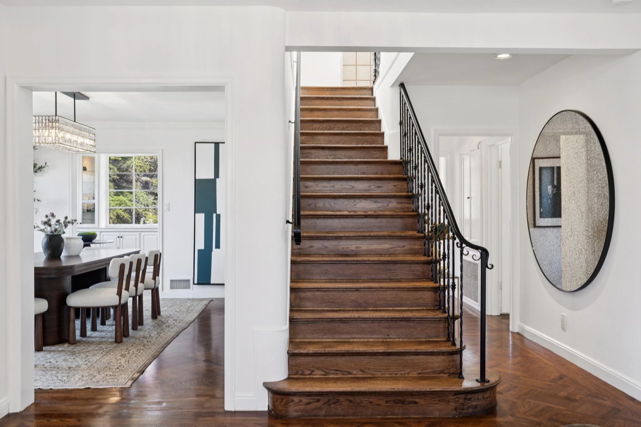 Interior view of a home foyer showing a staircase with wooden steps and black metal railing, a dining area with a large dark wood table and white upholstered chairs, a large window letting in natural light, and decorative elements like a circular mir