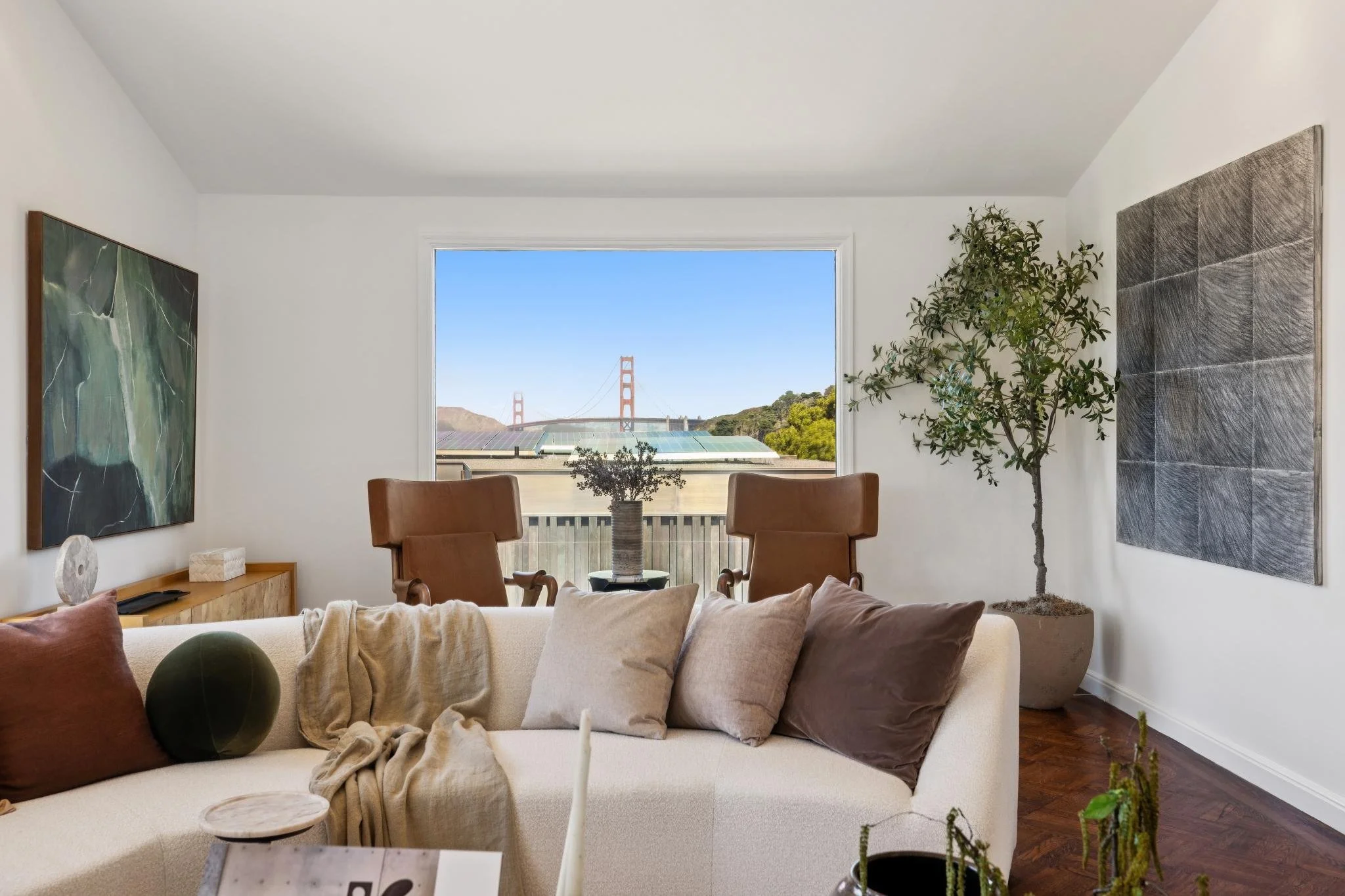 Living room with large window overlooking the Golden Gate Bridge, white walls, and modern art. Contains beige sofa with brown and cream pillows, wooden floor, green potted plant, and chairs.