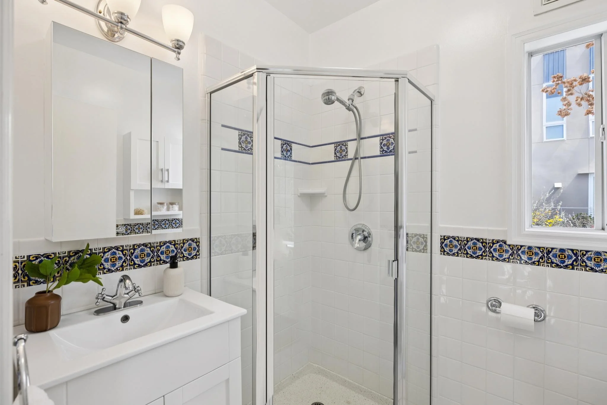 A bright bathroom featuring a shower with glass doors, a white sink with a cabinet, a mirror, and a window with a view of neighboring buildings.