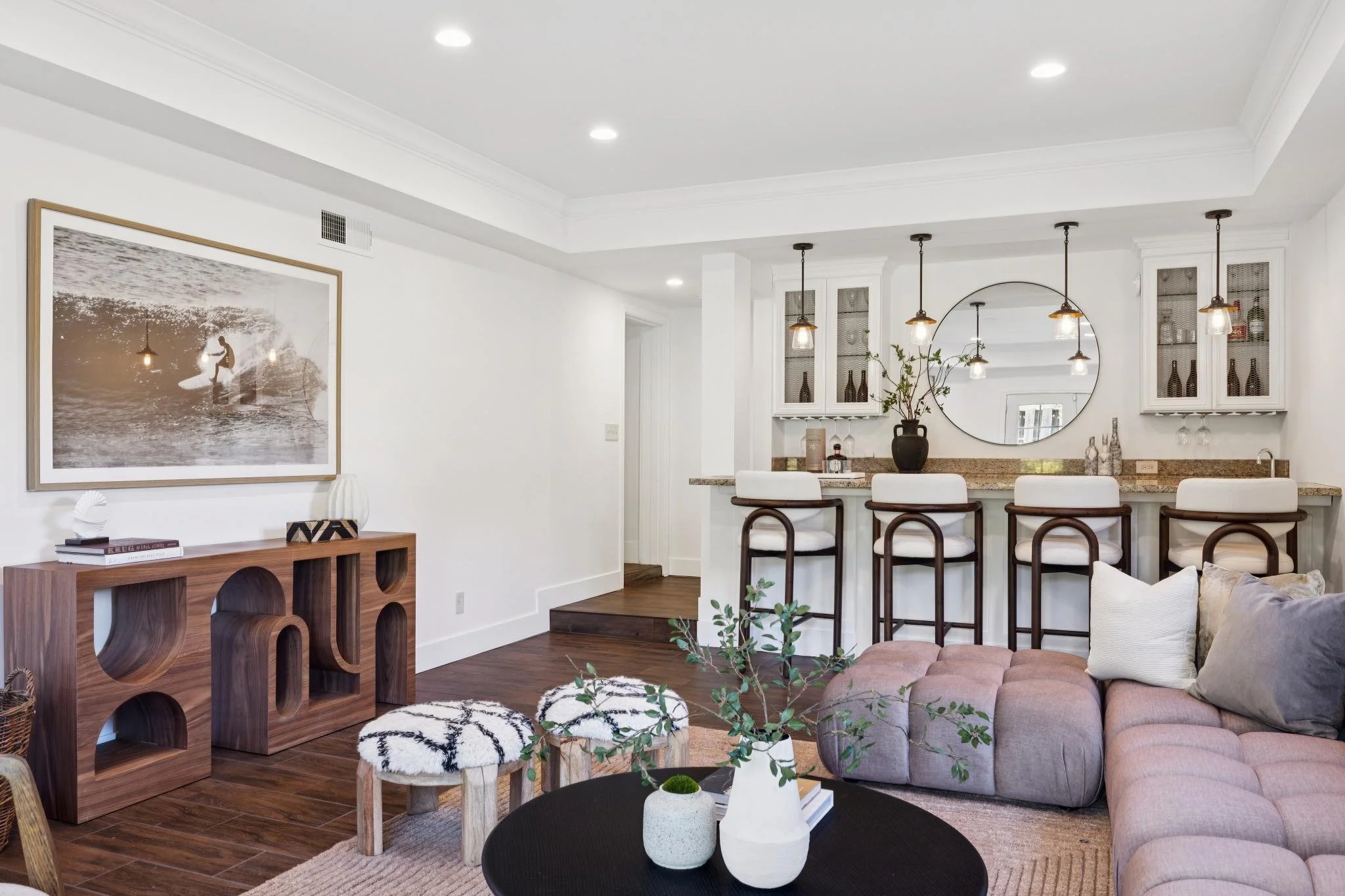 Living room with white walls, dark hardwood floors, and modern furniture including a pink sofa, black round coffee table with white vases, wooden sideboard, and bar stools at a kitchen counter. Decor includes a large framed surfing photo, a round mir
