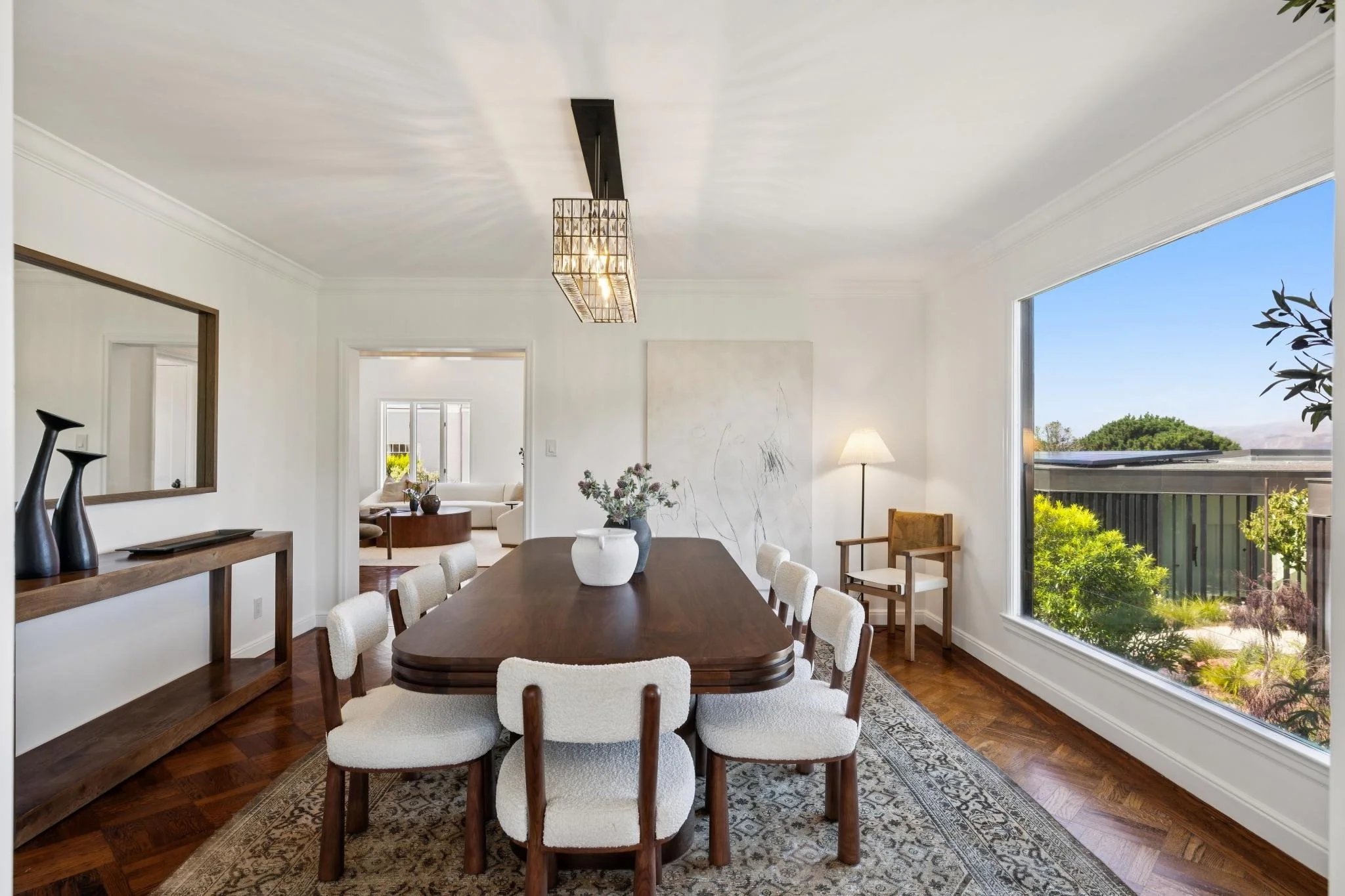 A bright dining room with a large window showing greenery outside. The room has a wooden dining table surrounded by ten white upholstered chairs, a wooden sideboard with decorative vases, and a modern chandelier hanging from the ceiling. A vase with 
