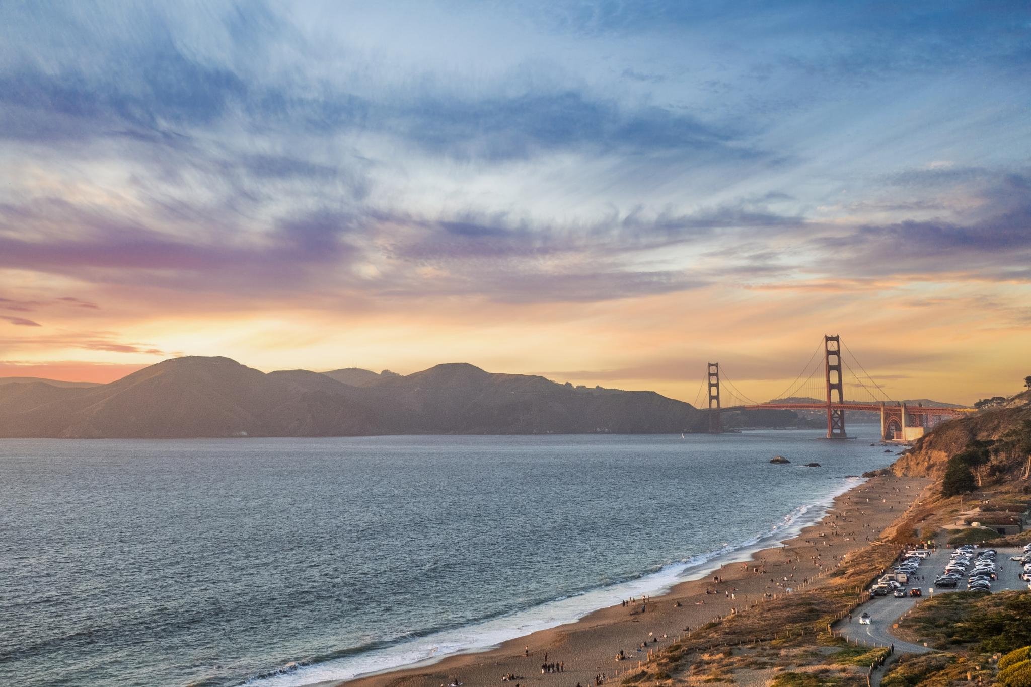 Golden Gate Bridge during sunset with hills in the background and people on the beach.
