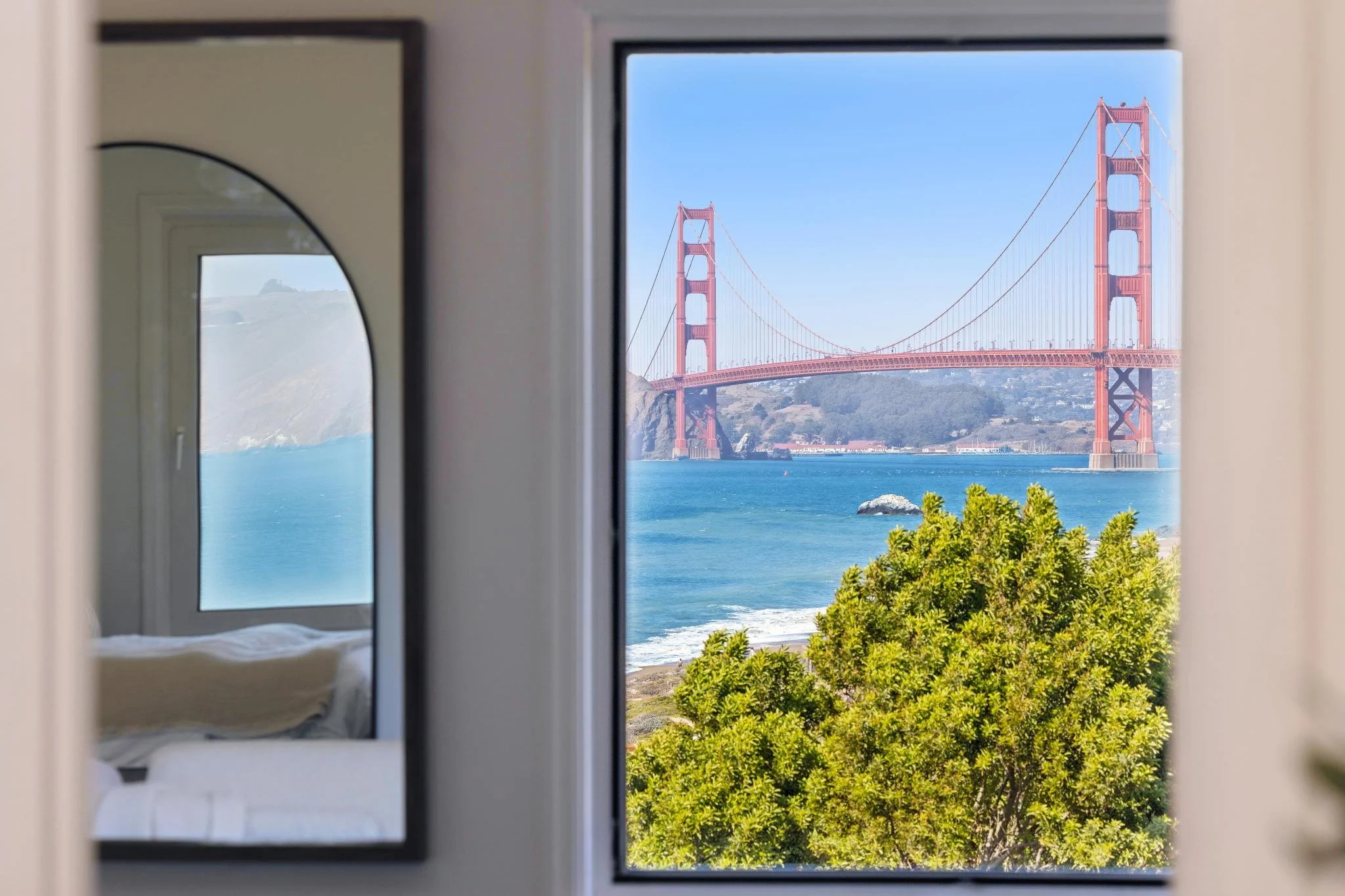 View of the Golden Gate Bridge through a window, with blue sky and green foliage in the foreground.