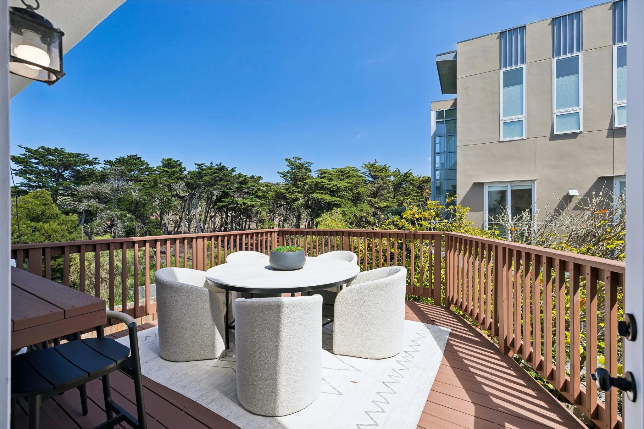 Patio with white outdoor dining table and four white chairs, red wooden railing, and trees in the background under a blue sky with some clouds.