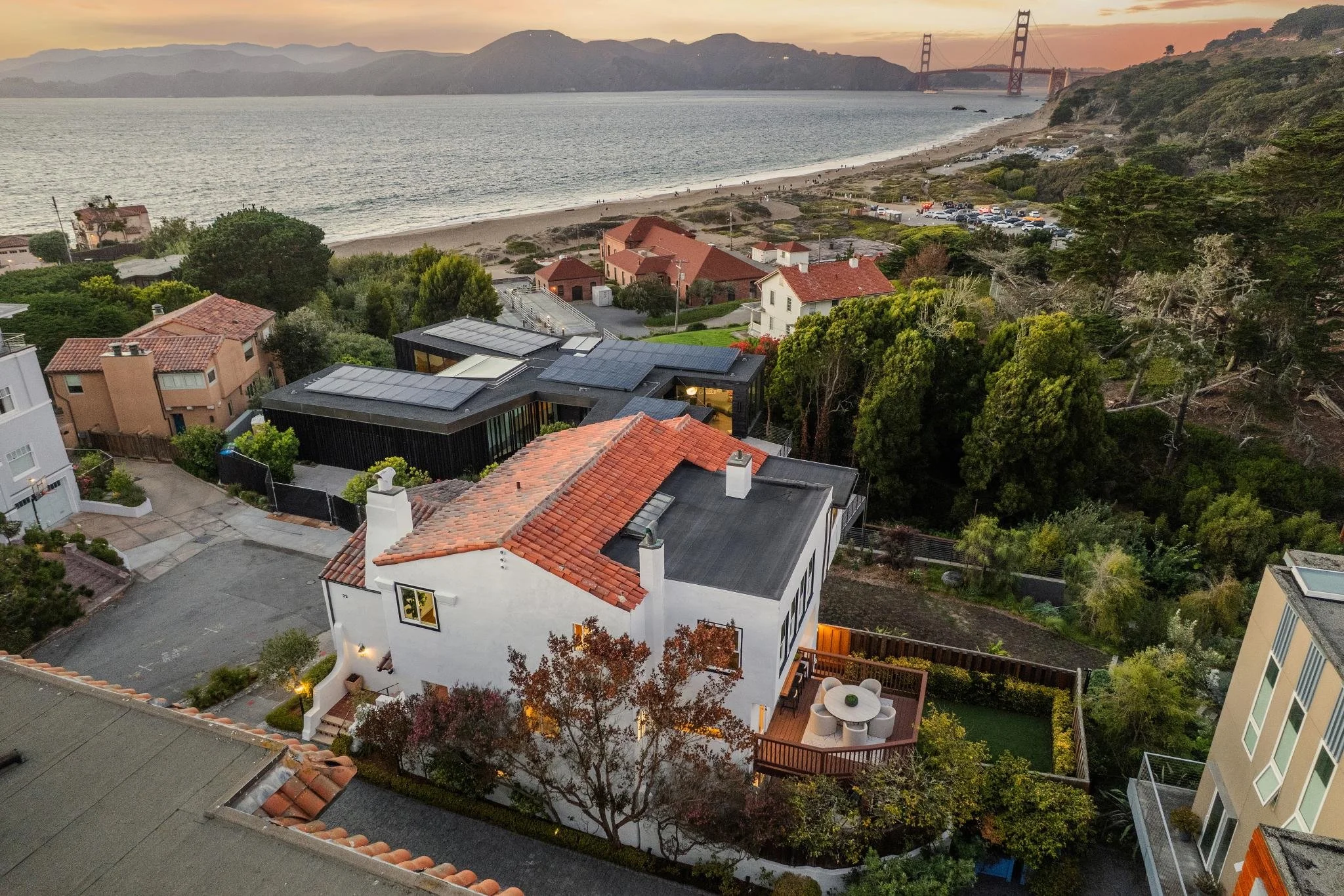 A scenic view of residential houses and buildings near a beach during sunset, with the Golden Gate Bridge visible in the background, and green trees surrounding the area.
