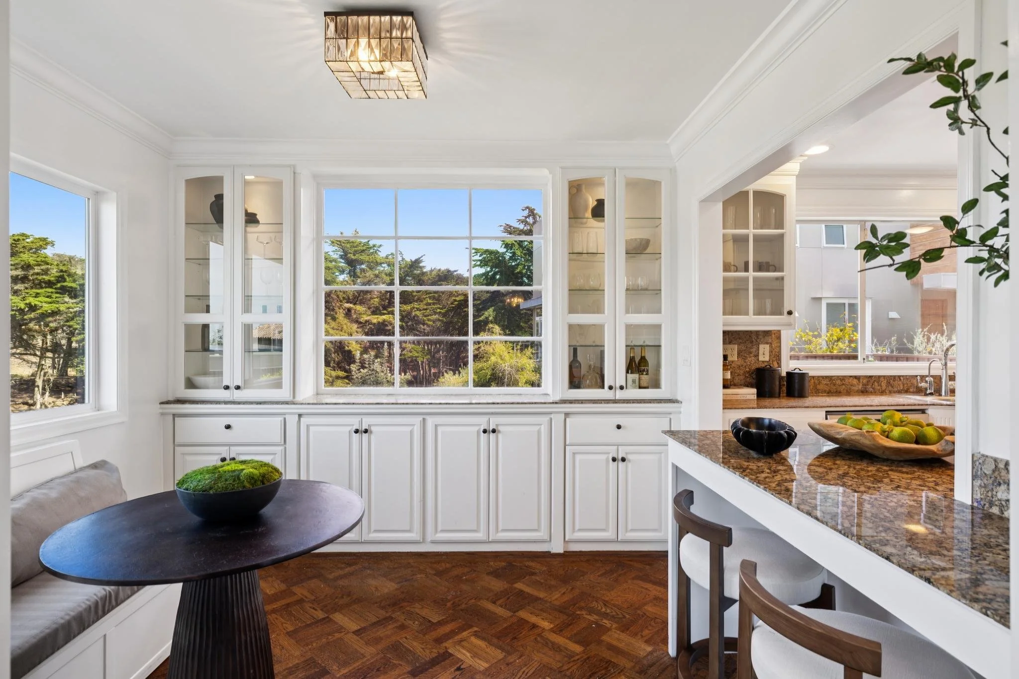 Bright kitchen with large window, white cabinets, granite countertops, and a small dining area with a black round table and bench seating.