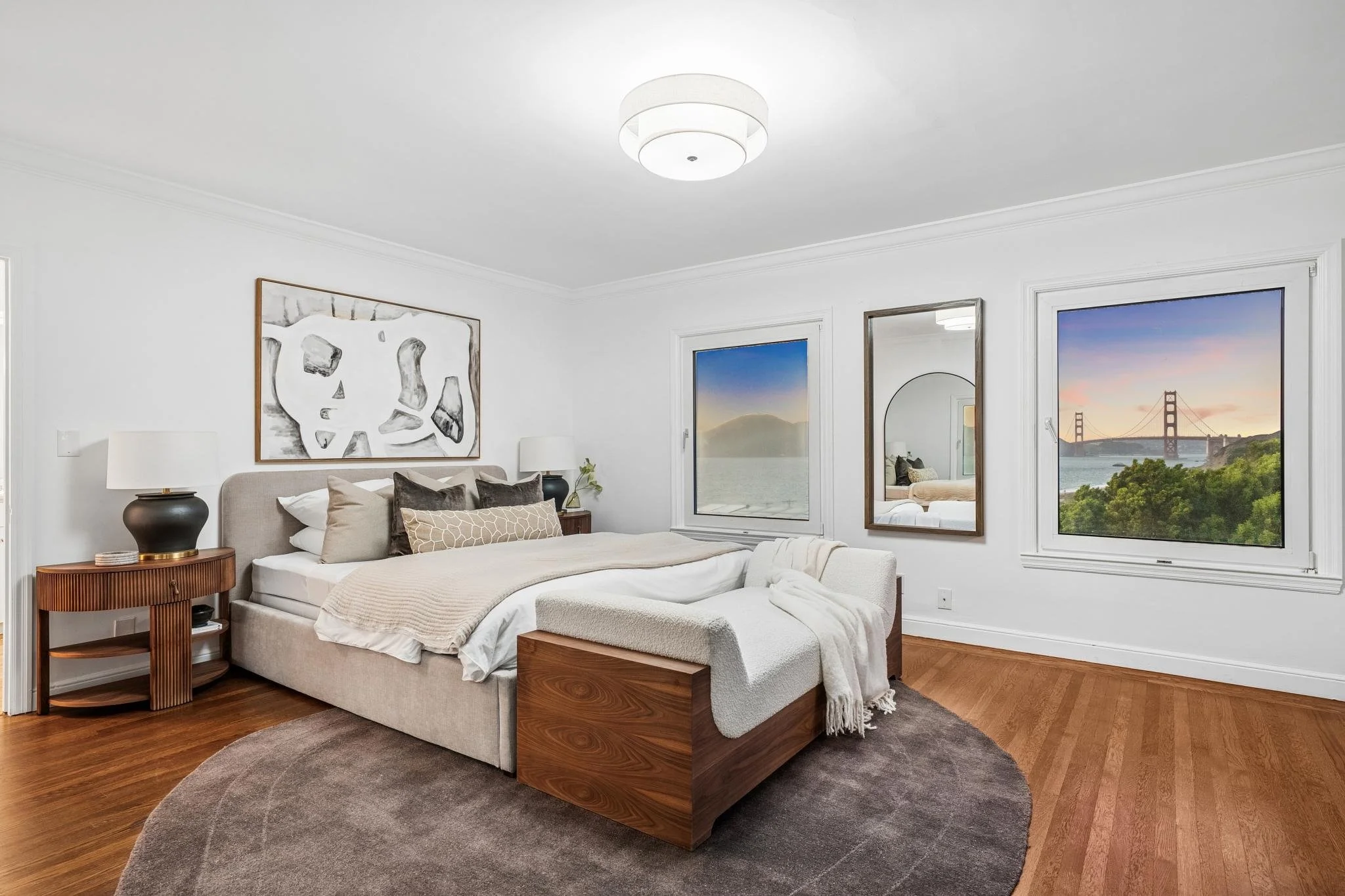 A bedroom with a beige bed, wooden nightstands, lamps, and artwork on the white walls. Two windows show a view of a bridge and a landscape at sunset.