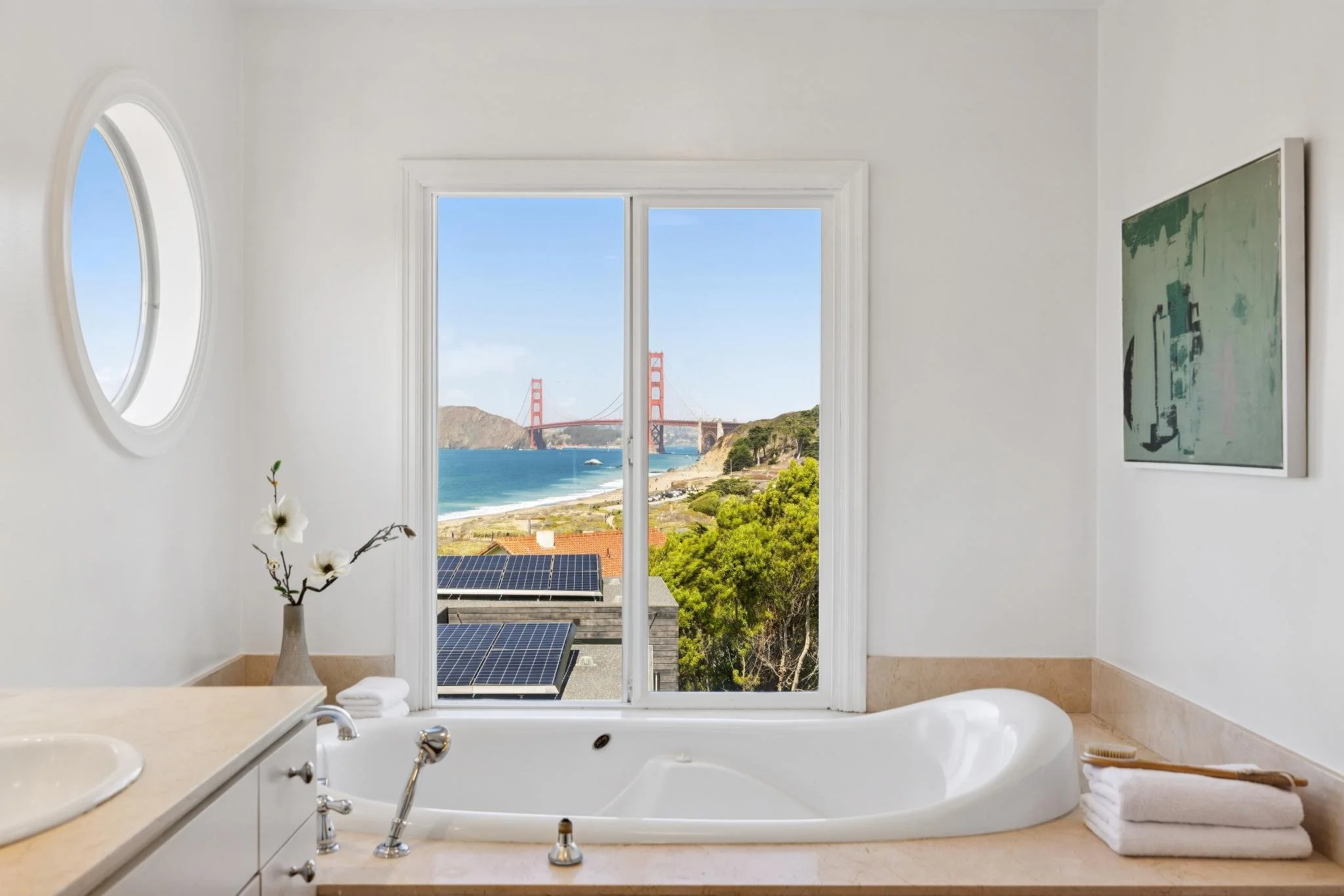 A bathroom with a large window and a view of the Golden Gate Bridge in San Francisco. The bathroom has a bathtub, a flower vase with white flowers, folded towels, and a bathroom countertop.