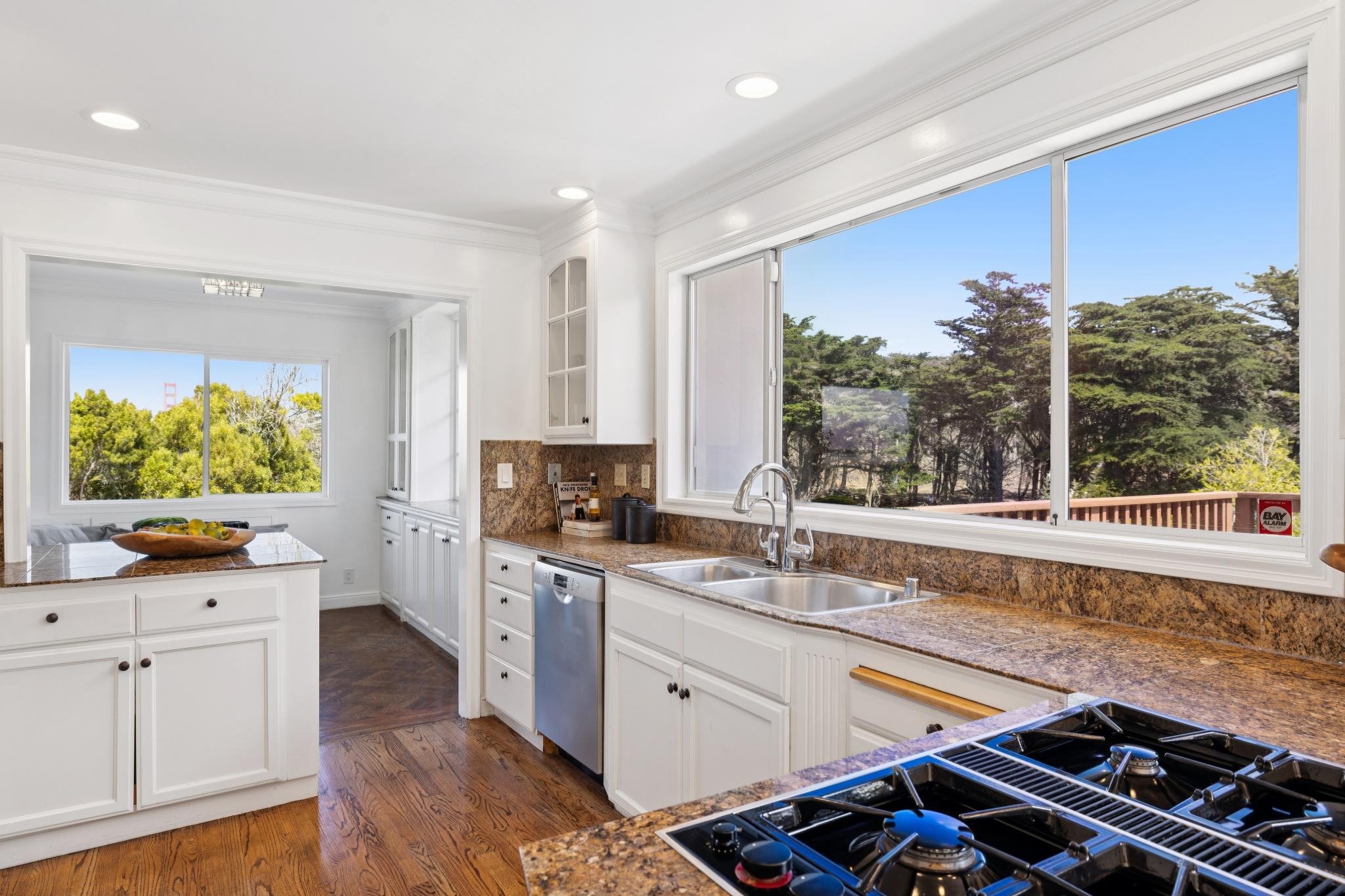 Bright kitchen with large windows showing green trees outside, white cabinets, brown granite countertops, and a cooktop with burners.