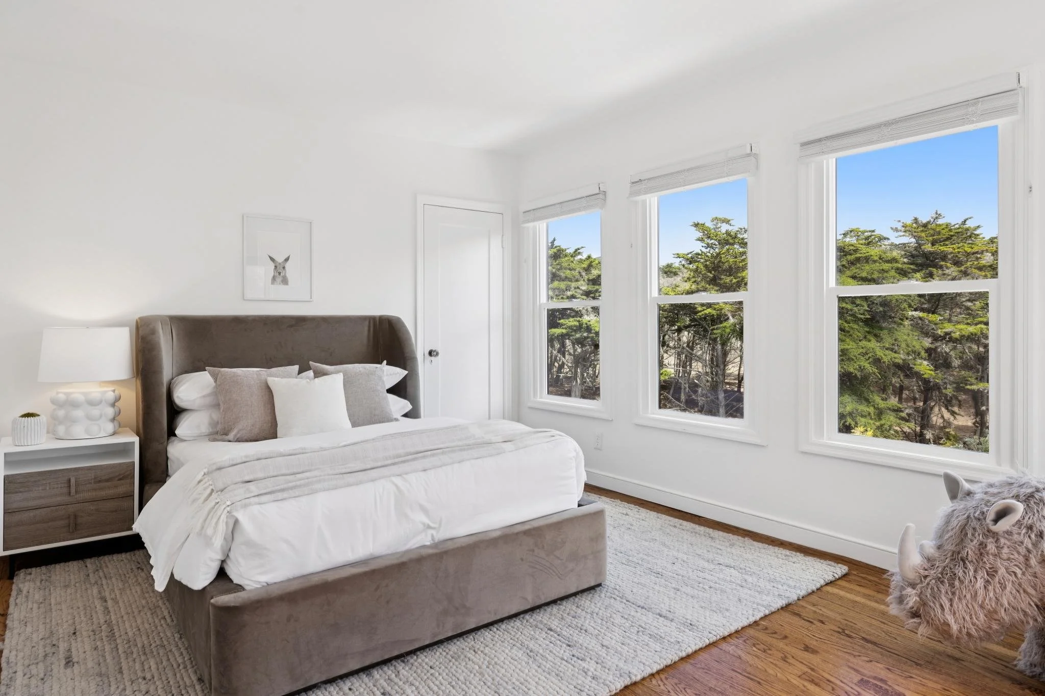 Bright bedroom with white walls, large windows, beige bed with white linens, and furry dog lying on the floor.