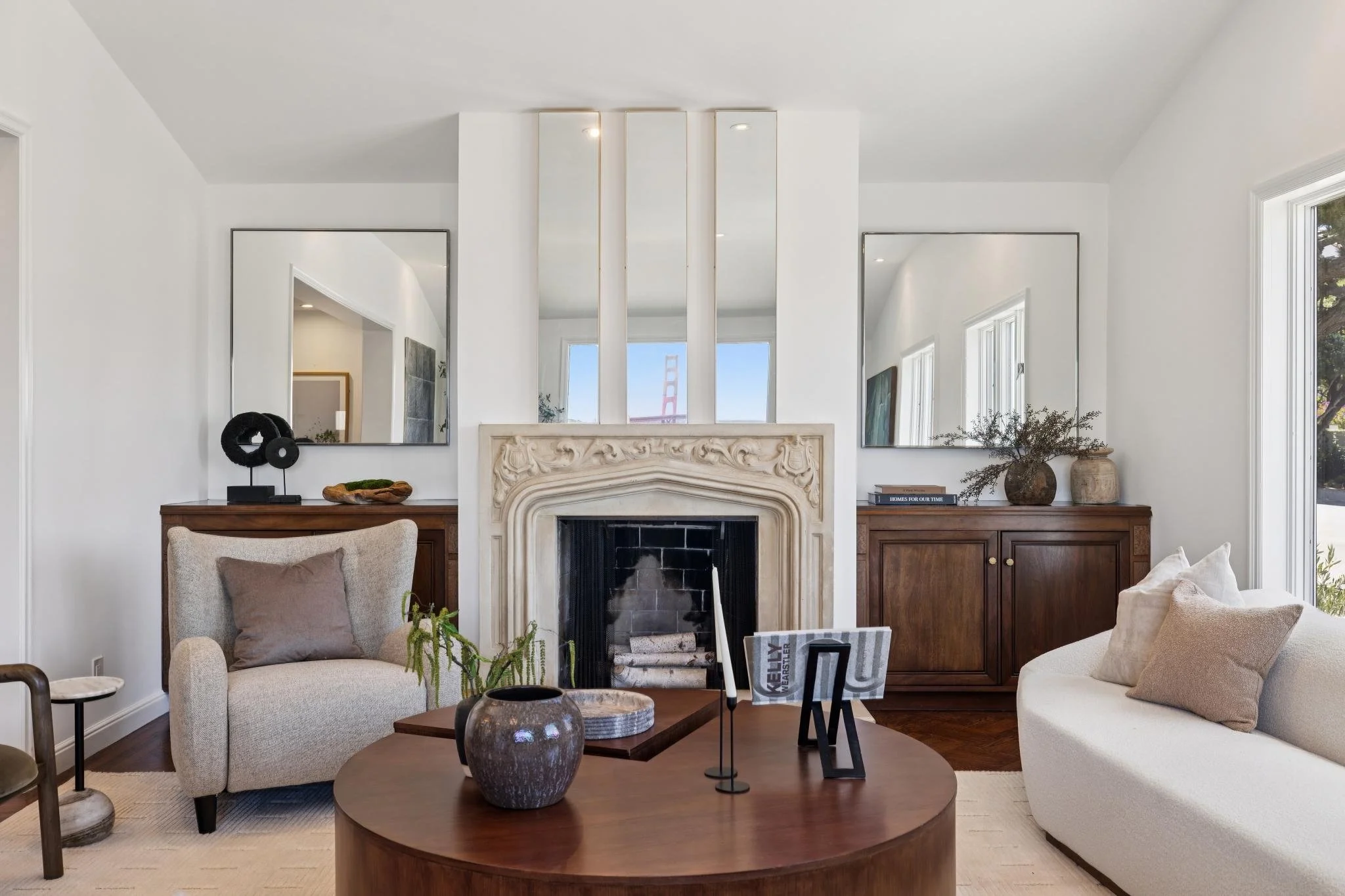 Living room with beige armchair, white sofa, wooden coffee table with decorative vase, large mirror above fireplace, dark wooden sideboards, and large windows revealing trees outside.