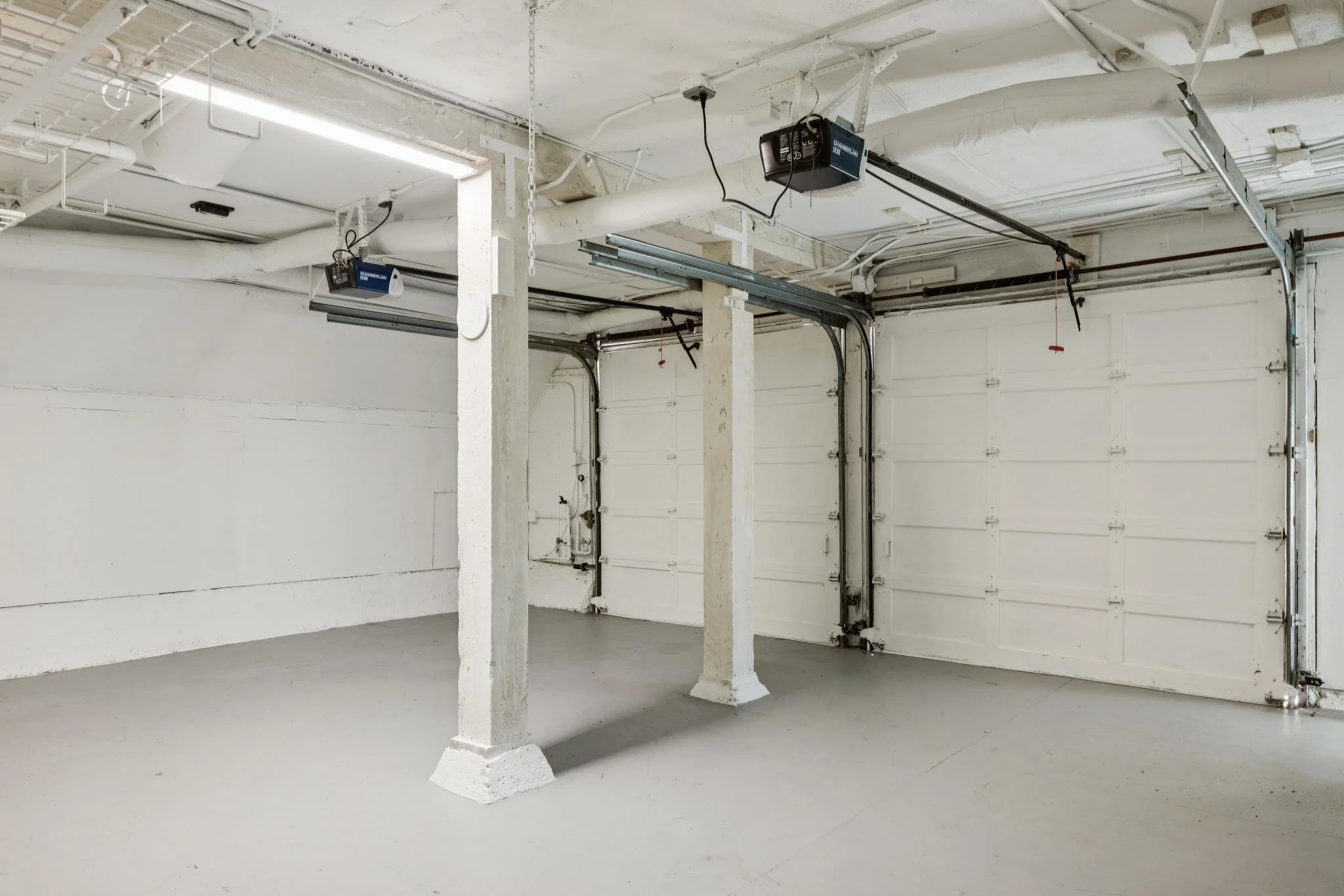 Empty garage space with white walls, concrete flooring, a white garage door, and exposed piping and electrical wiring on the ceiling.
