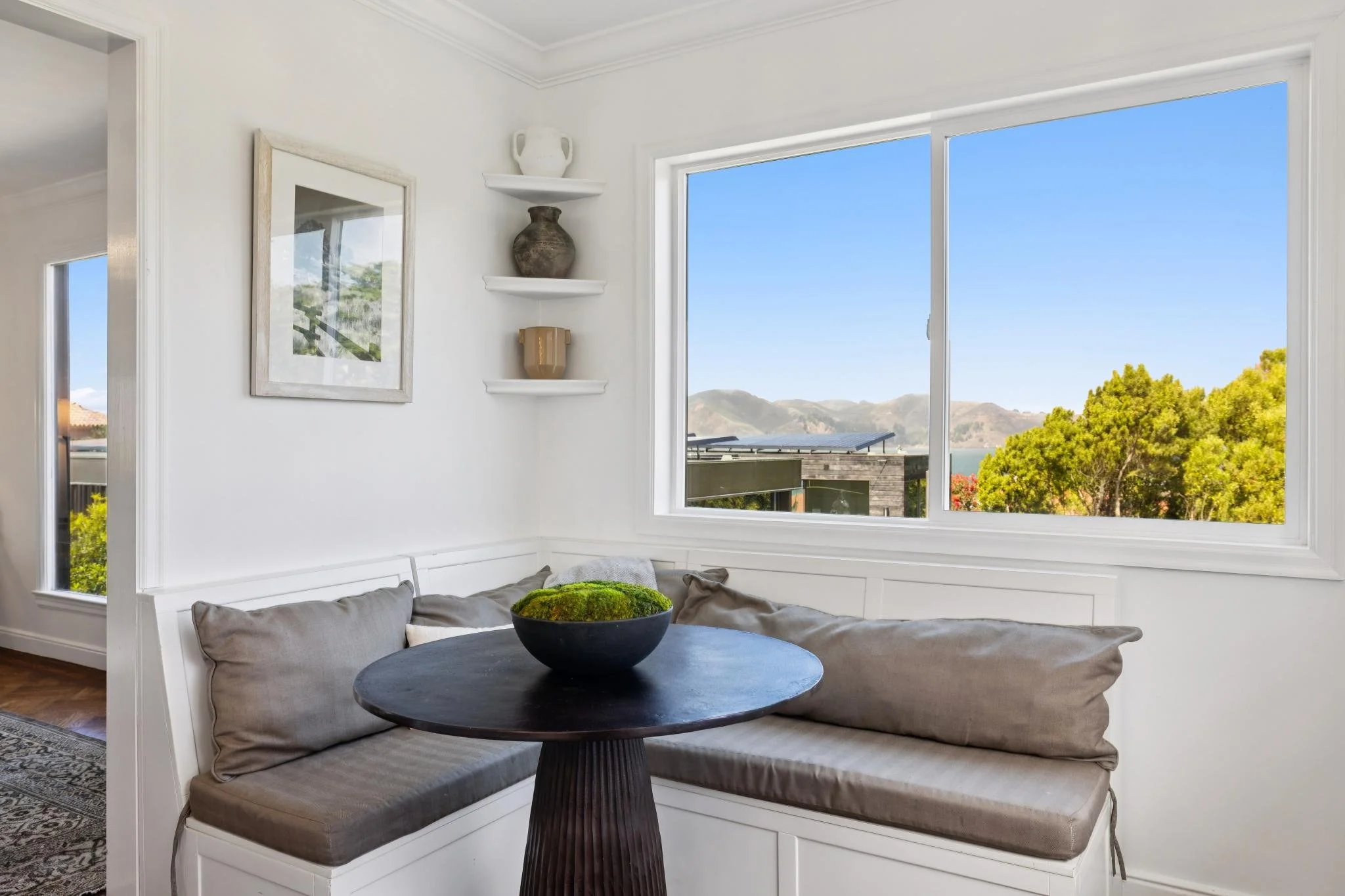 Living room with large window showing a mountain view, white walls, a framed picture, three shelves with vases, a round black table with a bowl of moss, and a built-in bench with gray cushions.