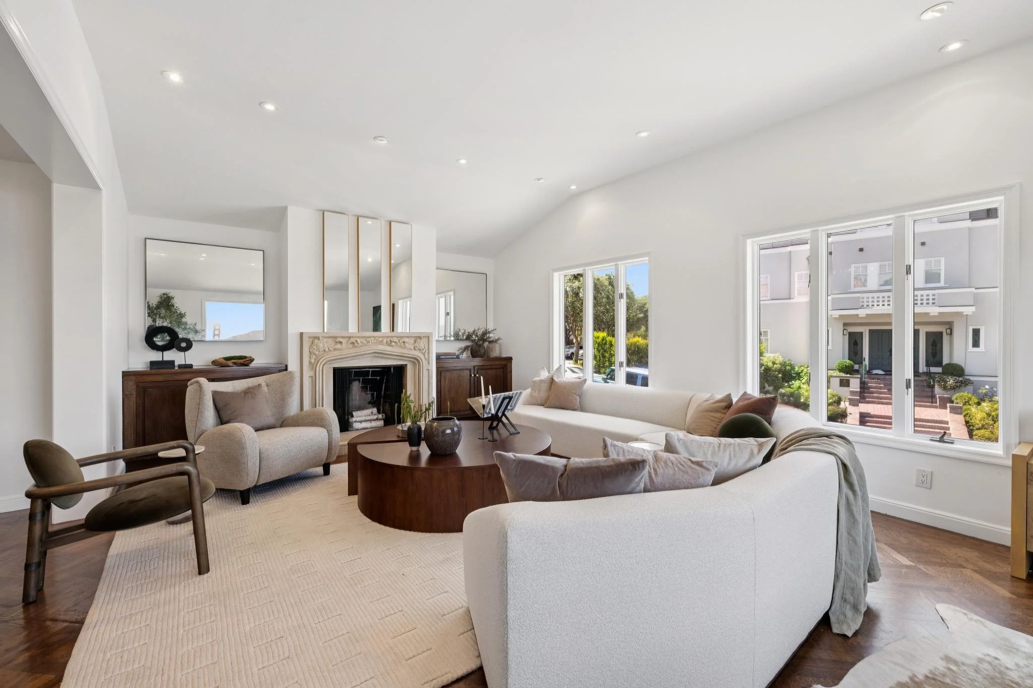 Bright living room with white walls and large windows, featuring a fireplace, beige sofas, a round wooden coffee table, and decorative items.