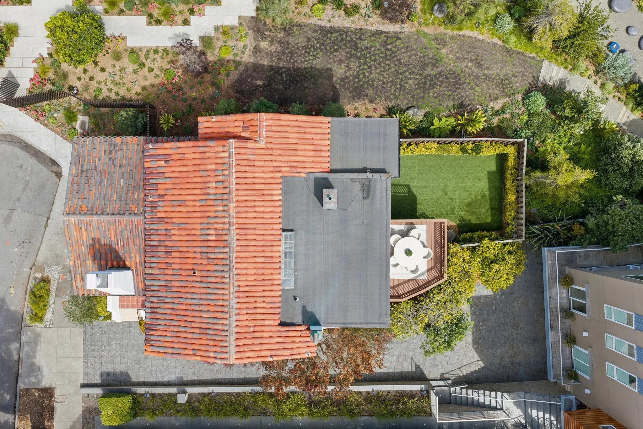 An aerial view of a residential backyard with a house with a red-tiled roof, a concrete patio, a lawn, and garden beds with various plants and trees, surrounded by a wooden fence.