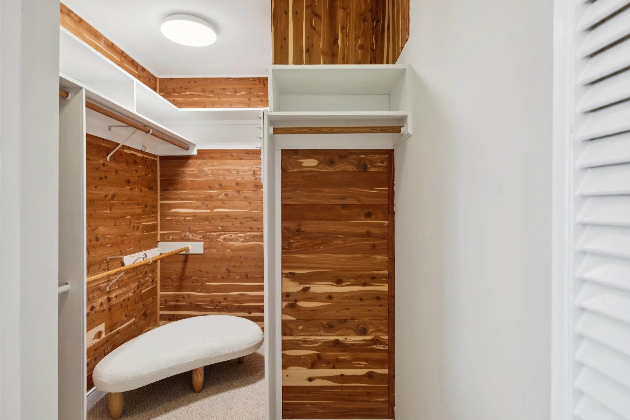 A walk-in closet with wooden walls, white shelving and closet rods, a beige cushioned bench, and white louvered shutters on the right side.