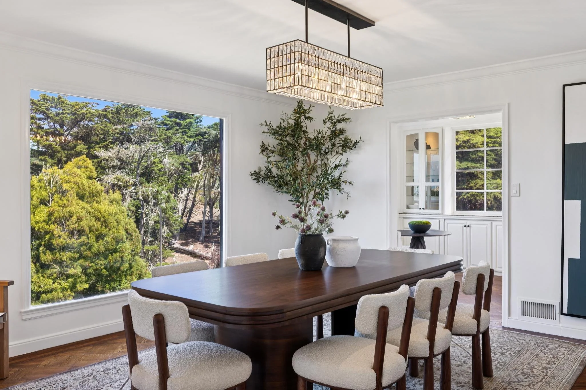 Dining room with large windows showing trees outside, a dark wood table with six cream-colored upholstered chairs, decorative vases and plant on the table, white cabinetry with glass doors, and a modern chandelier hanging from the ceiling.