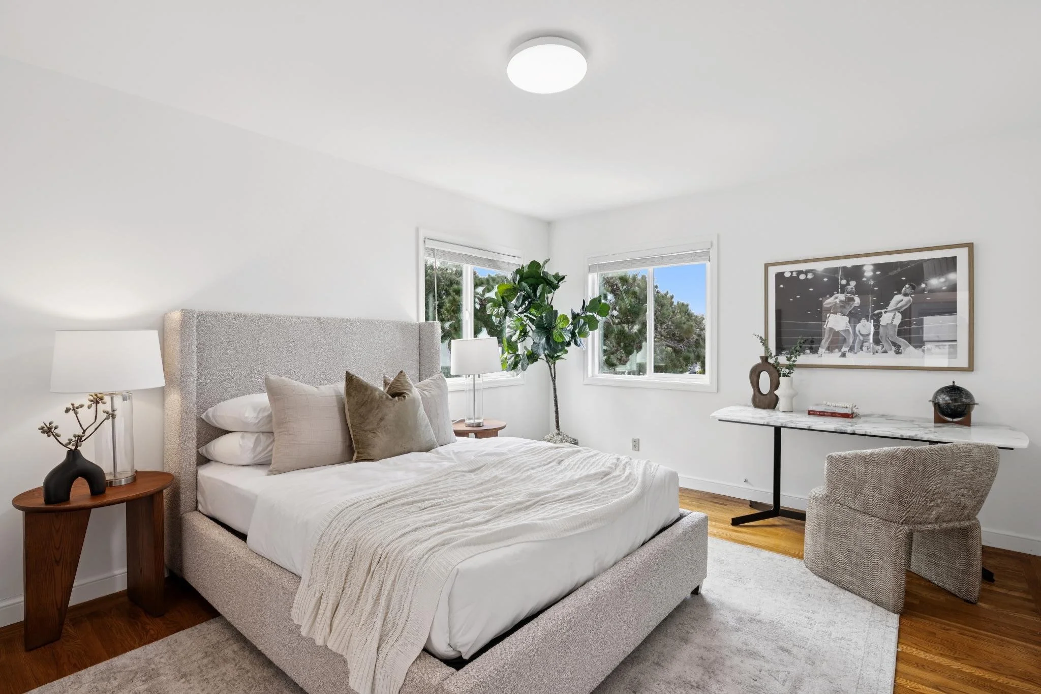 A modern bedroom with a neutral color palette, featuring a bed with white bedding and pillows, a wooden nightstand with a black decorative sculpture and lamp, a large potted plant near two windows, a framed black and white boxing photograph on the wa