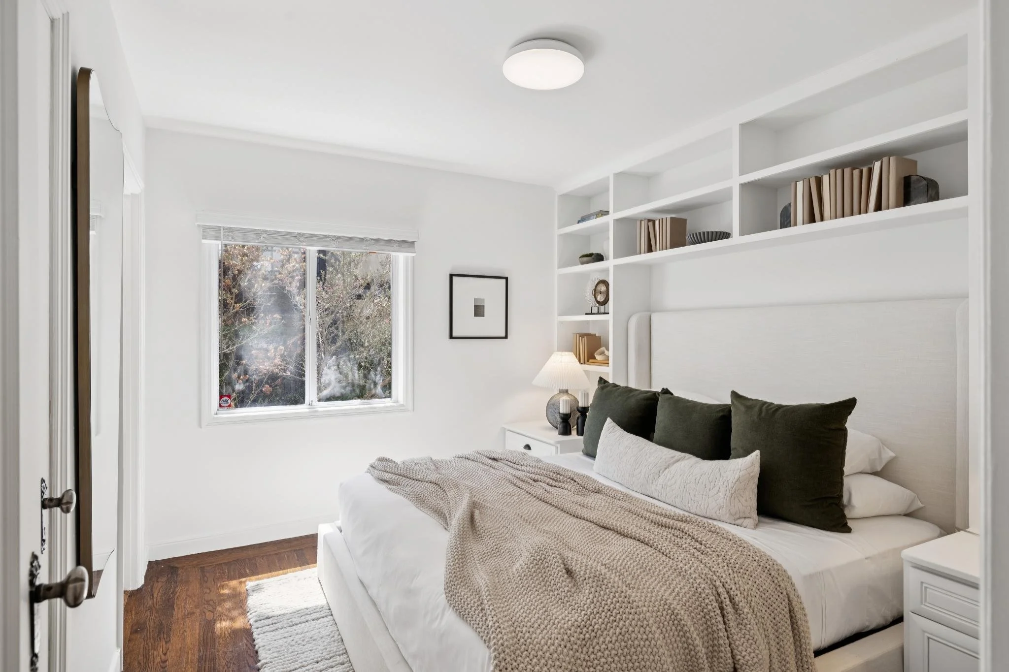 A bright bedroom with white walls, a window with blinds, a bed with pillows and a beige blanket, a white nightstand with a lamp and decorative items, and built-in white shelves with books and decor.
