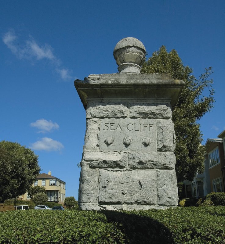 A stone monument engraved with "SEA CLIFF" and three shell carvings, situated on a grassy area with trees and residential buildings in the background under a blue sky.
