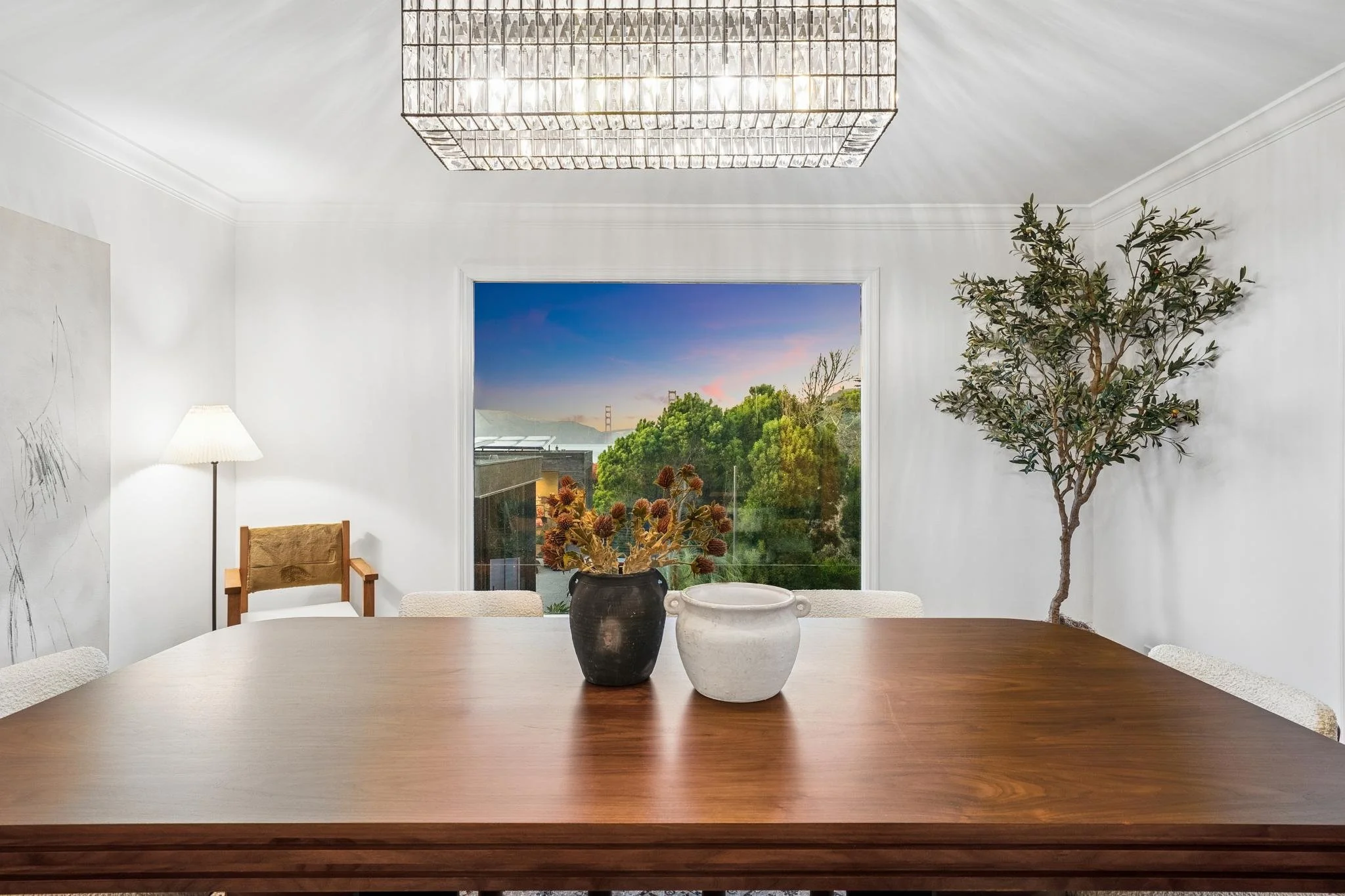 A dining room with a wooden table, decorative vases, a potted tree, a window showing a view of trees and the Golden Gate Bridge at sunset, and a modern chandelier hanging from the ceiling.