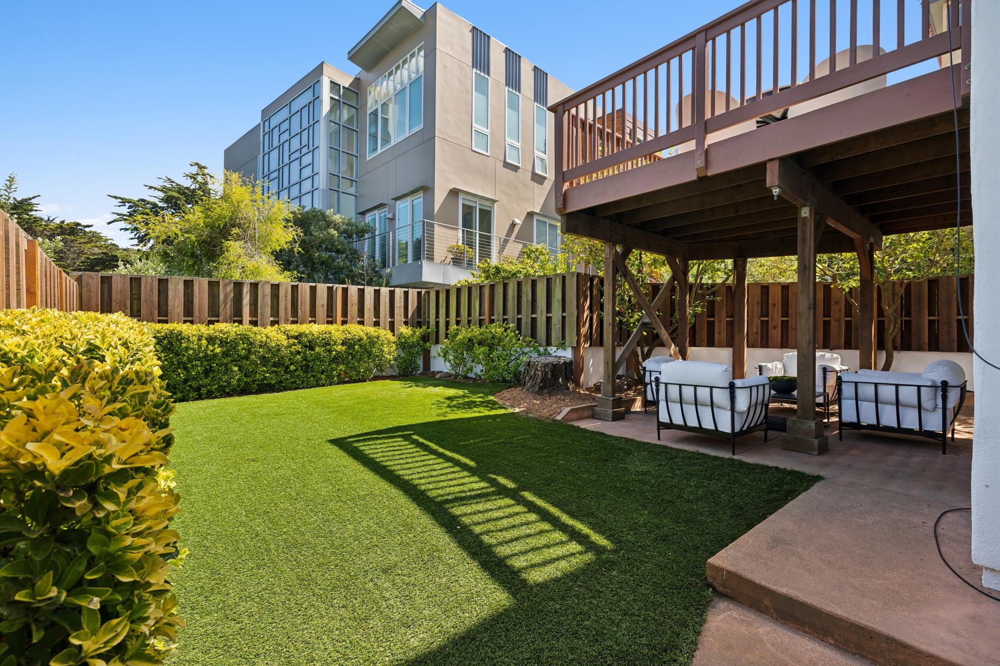 A backyard with artificial grass, a wooden privacy fence, lush green bushes, trees, and an outdoor seating area under a wooden pergola with cushioned chairs. In the background, there's a modern multi-story apartment building with large windows and a 